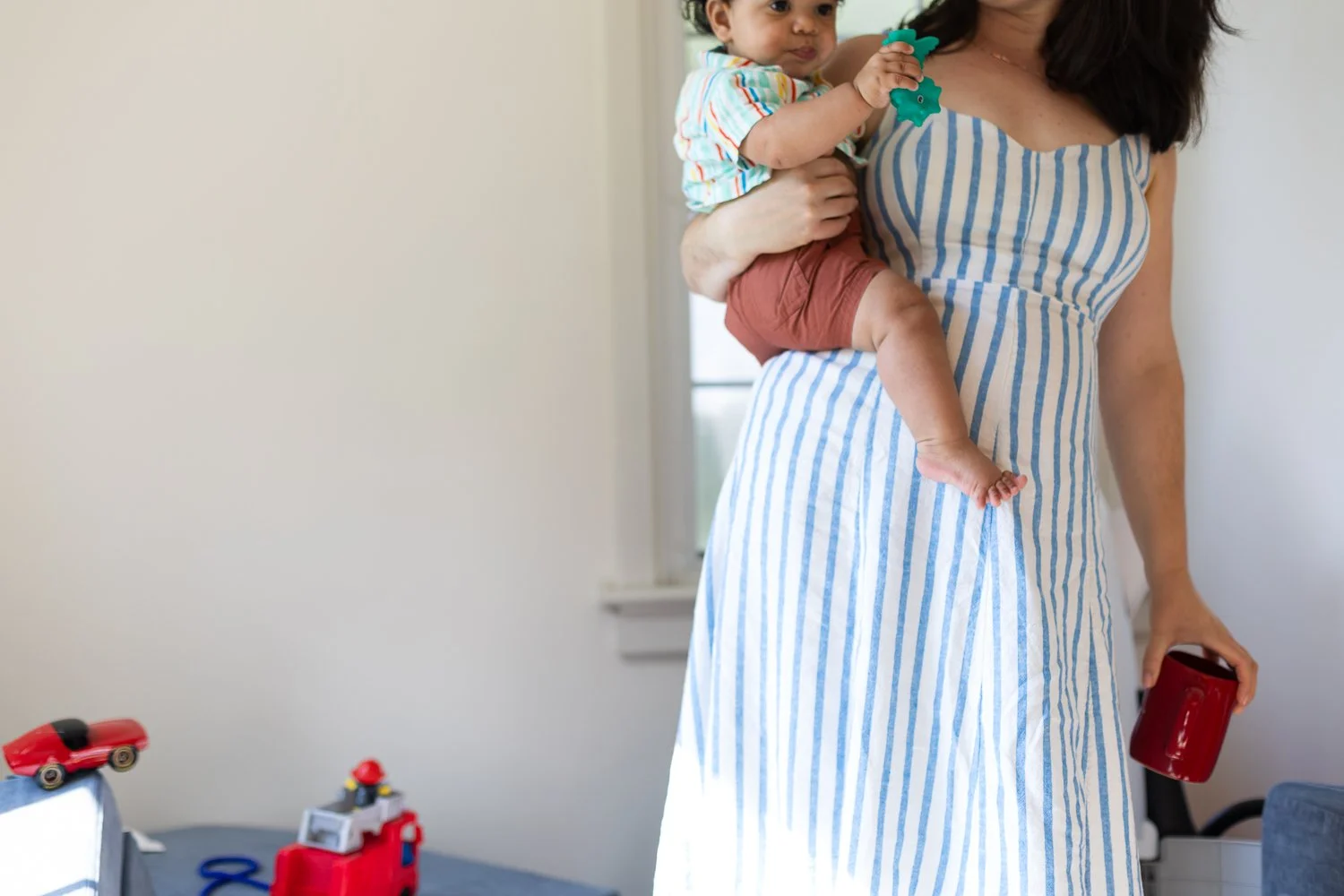 A mom carries her infant son on one hip and a coffee cup with her other hand. He is carrying a chew toy.