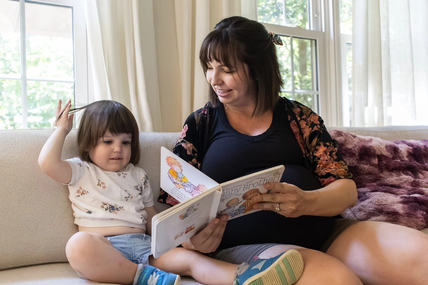A pregnant woman and a young girl sitting on a beige couch, reading a children's book called 'Baby Brother,' with a window and curtains in the background.