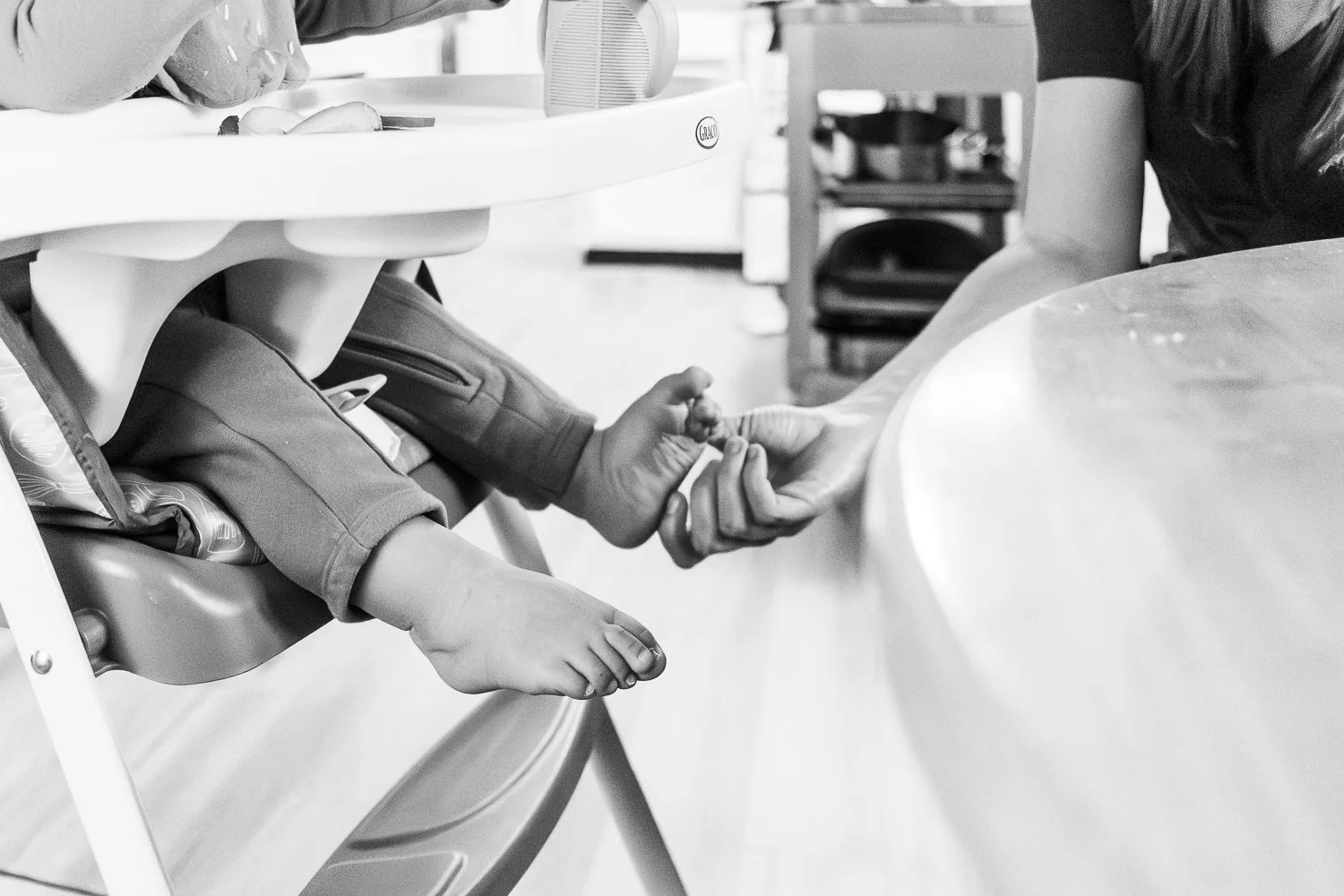 A mom tickles her baby's foot while they sit in their high chair with a snack. The photo is black and white.