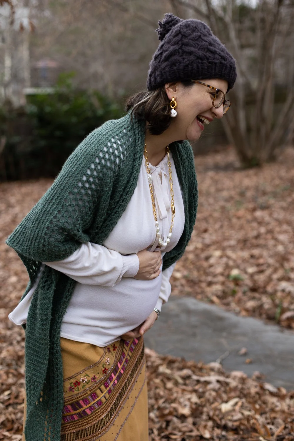 A pregnant woman in a winter hat and glasses laughing outdoors, wearing a green shawl, white blouse, and a patterned skirt, standing in her backyard with fallen leaves.