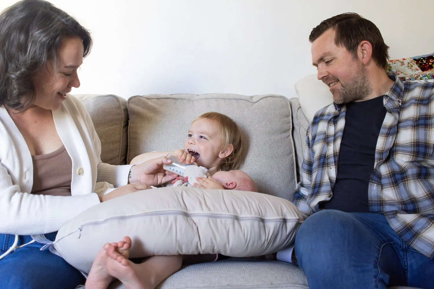 A little girl sits on the couch between her parents with her newborn sister in her lap. She gives the baby a bottle filled with milk and smiles joyfully at her parents while they help her support her sister's head.