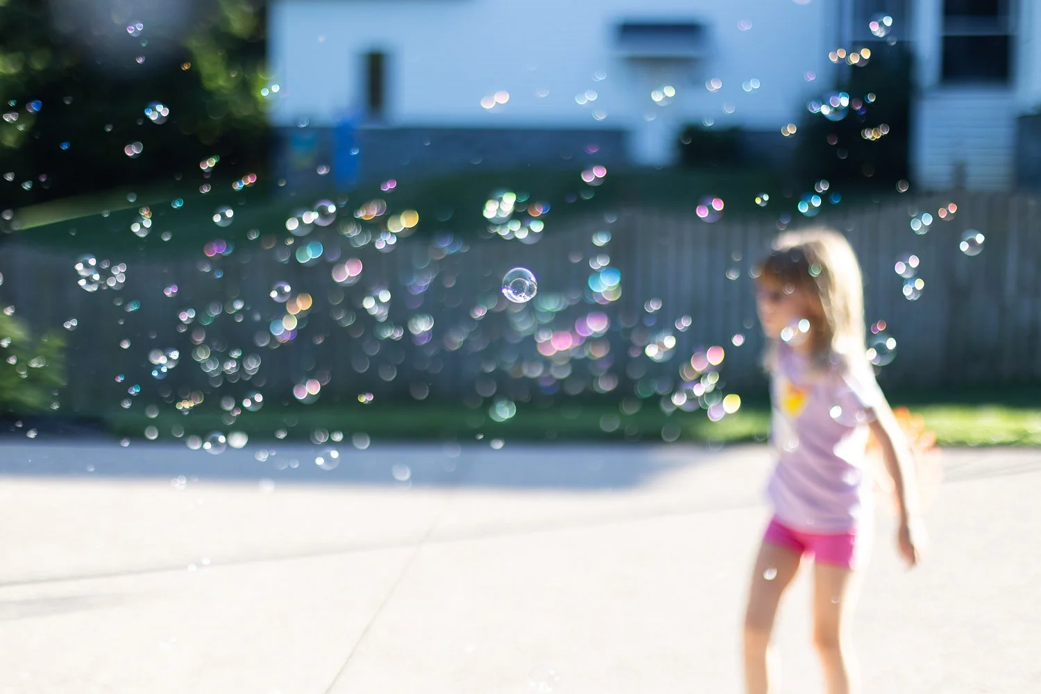 A young girl playing outside in a backyard, surrounded by floating soap bubbles with sunlight reflecting off them, with a house and fence in the background.