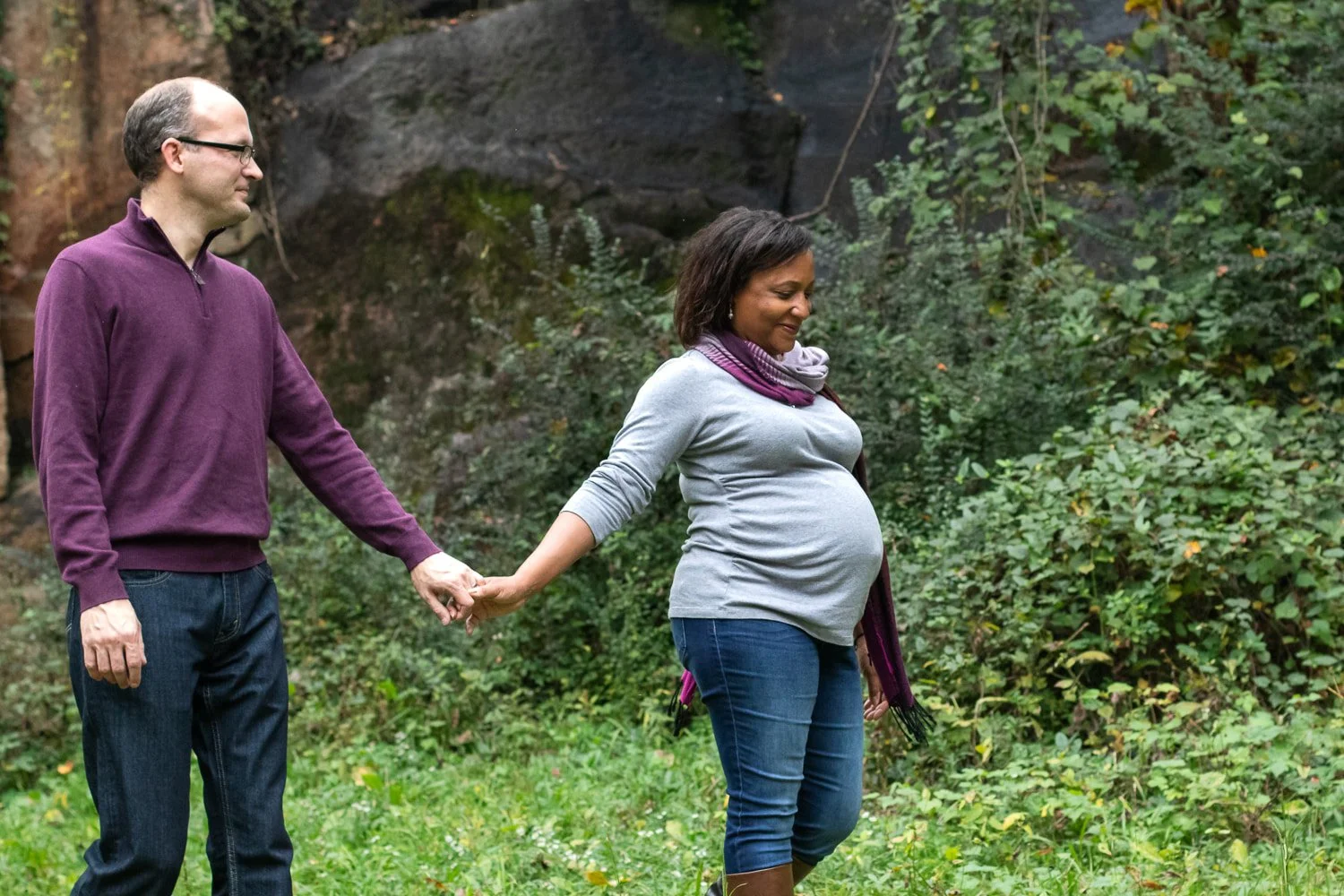 An expecting couple walk together while they hold hands. She is leading and smiling, and he is looking at her.