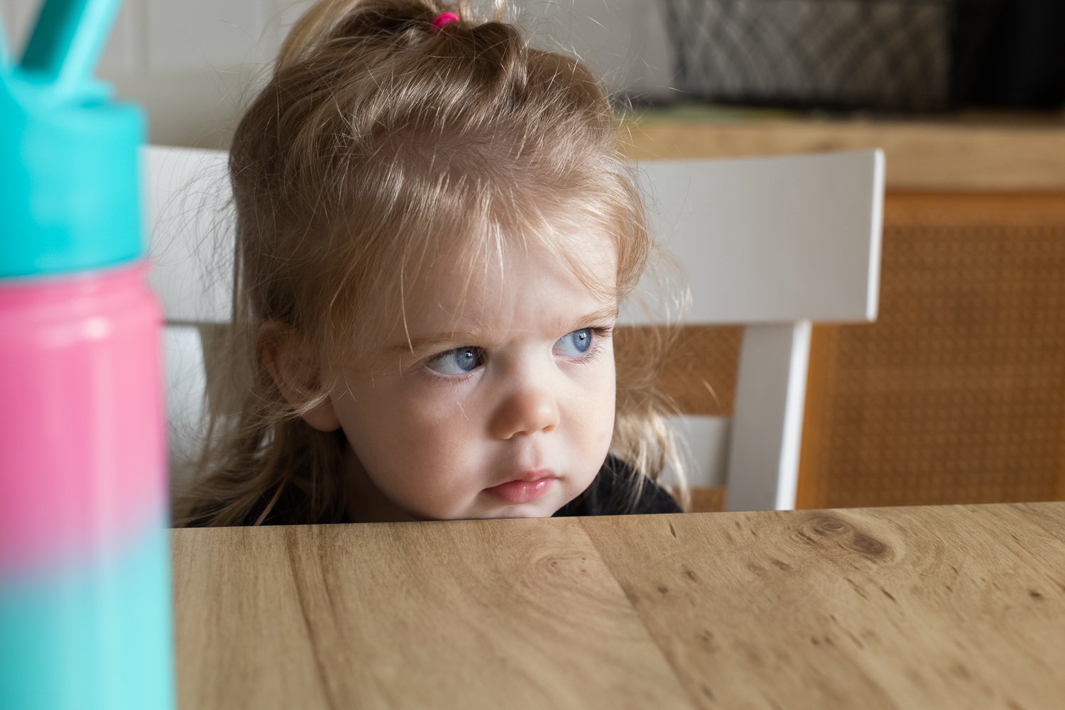 A little girl sits at a dining room table where her chin doesn't quite reach above the table level. Her bright blue eyes are looking off toward the window and her messy blonde hair is pulled back in a half ponytail.