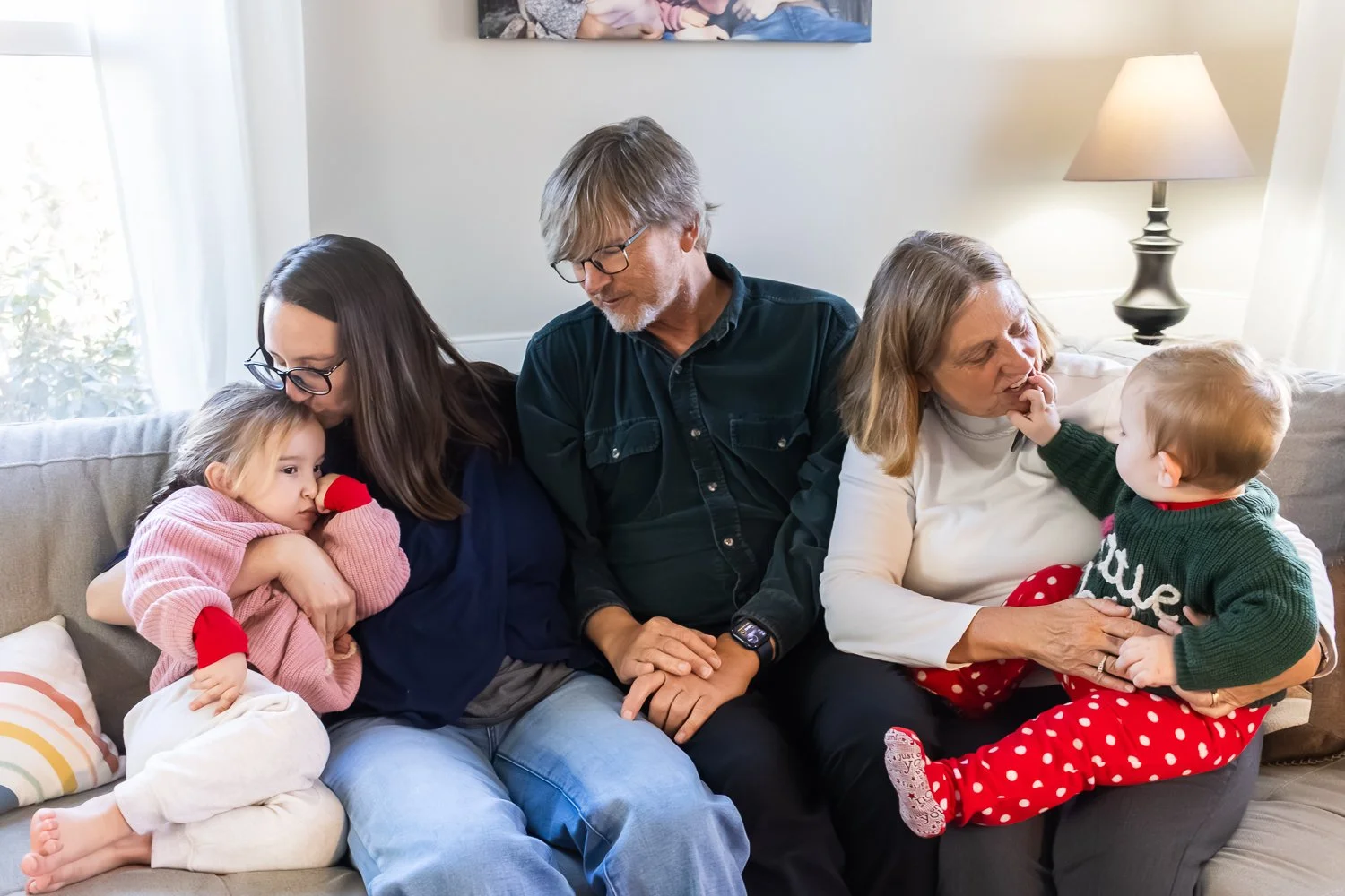 A mom, her two kids, and their grandparents sit on the couch together, no one is looking at the camera, they are all engaged with each other in various ways.