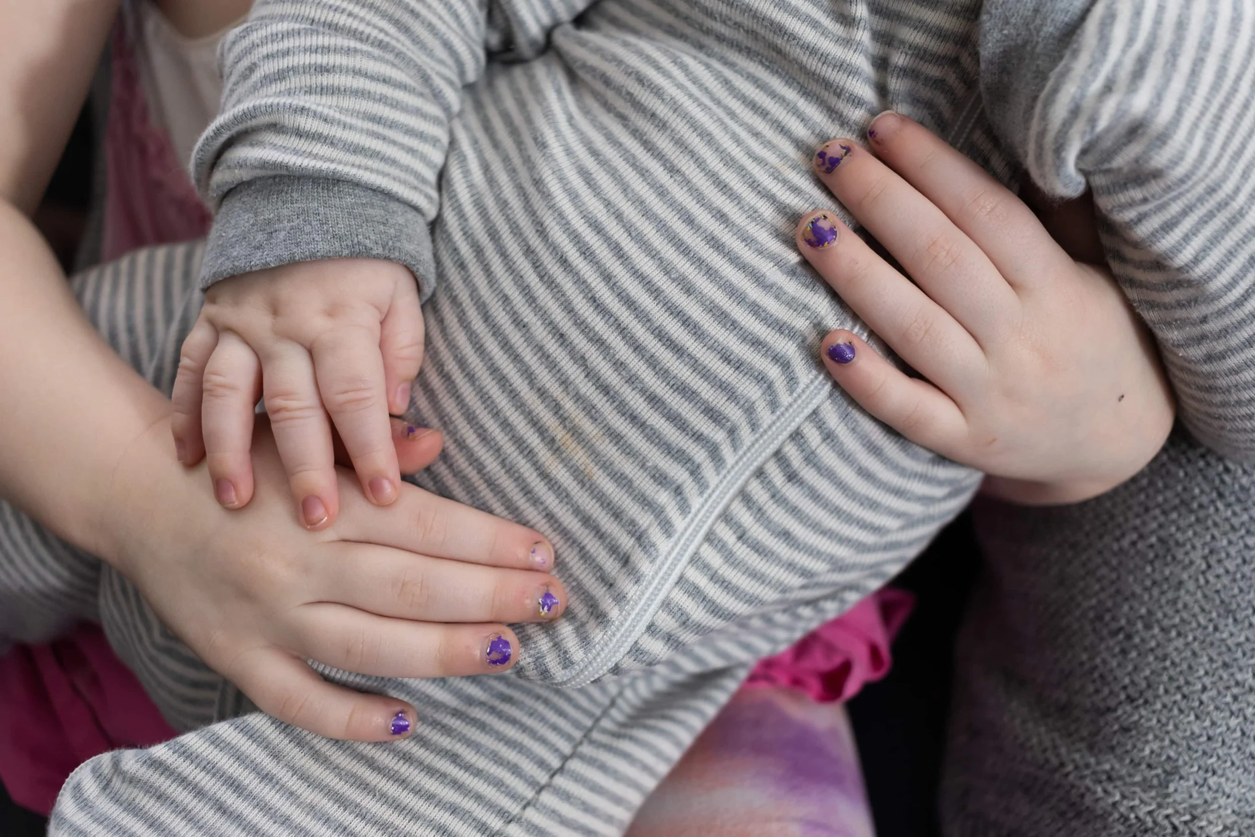 A baby's pajama-clad belly, with his toddler-sister's hands holding him. She has chipped purple nail polish and his chubby hand rests on one of hers.