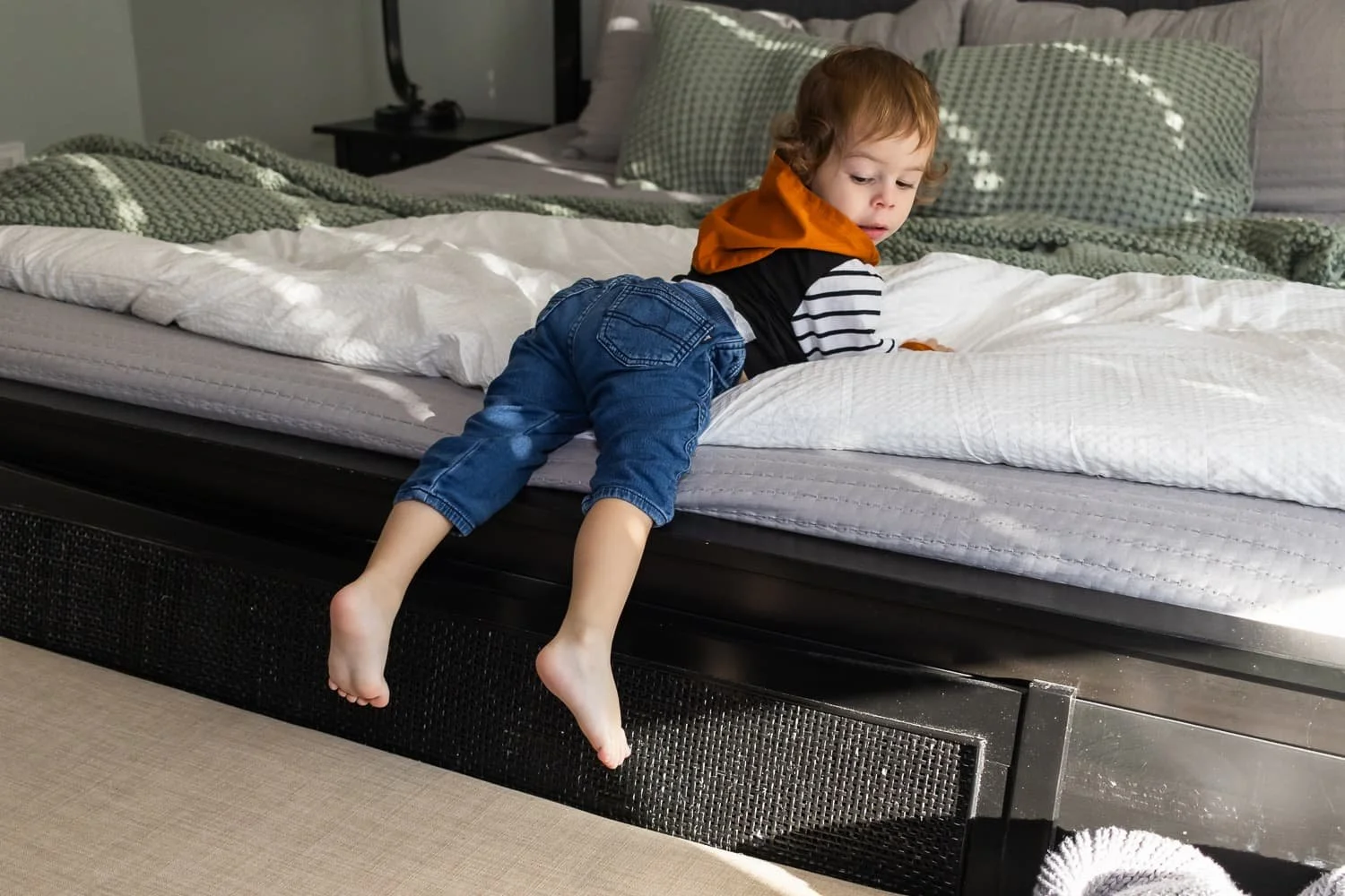 A young child with curly red hair wears a black and white striped shirt with an orange hood and denim shorts. The child is lying on their stomach on a bed, hanging off the edge with bare feet dangling toward the floor, looking down.