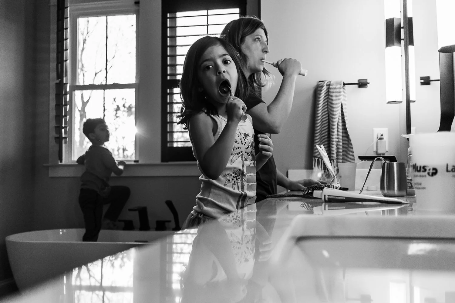 Mother and kids in the bathroom, little girl is staring at the camera with her toothbrush in her mouth while Mom stares at the mirror and her son climbs out of the bathtub (fully clothed) in the background.