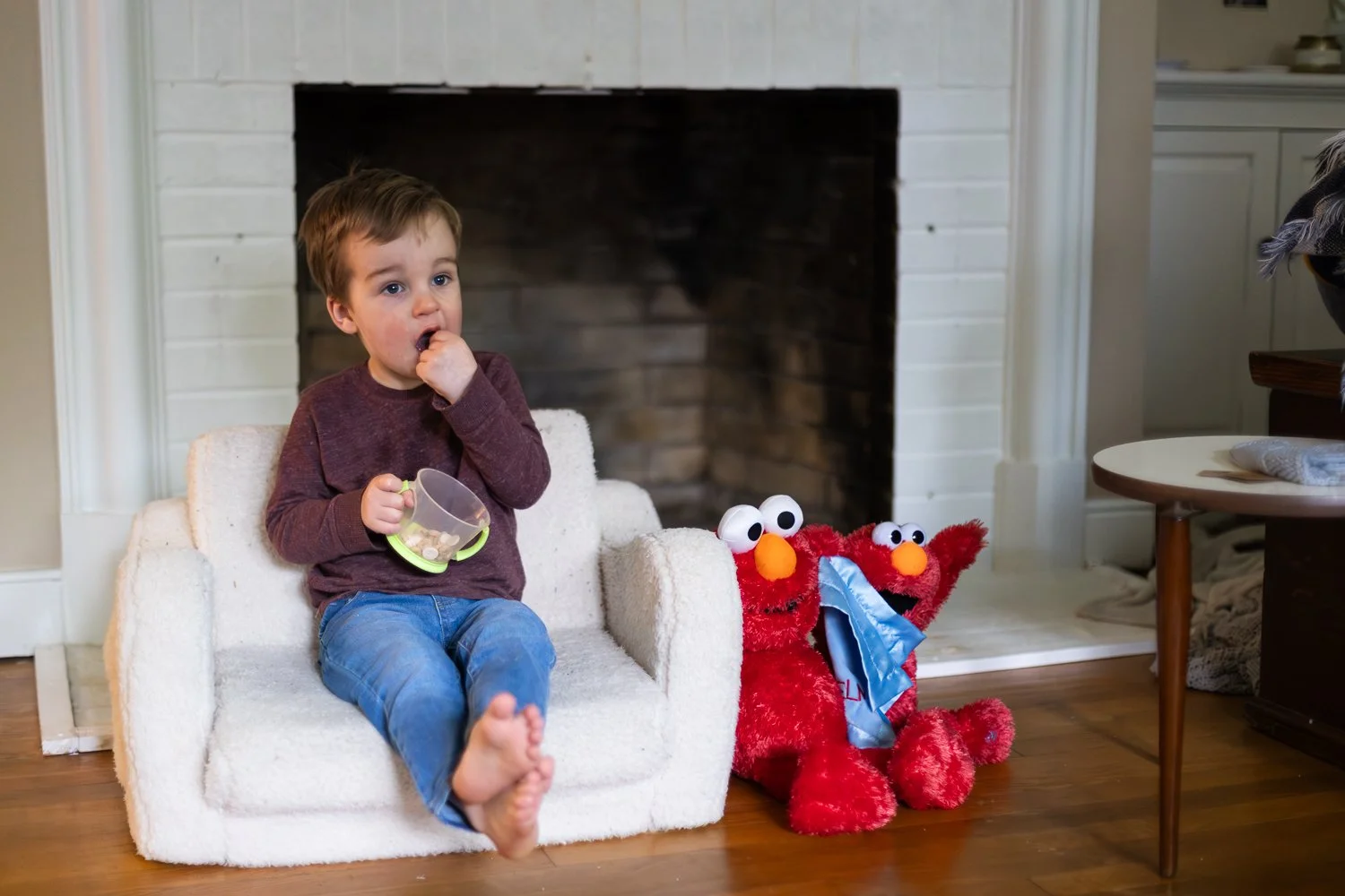 A toddler sits on a toddler-sized chair and eats snacks out of a cup in his living room. Two stuffed Elmos sit beside him.