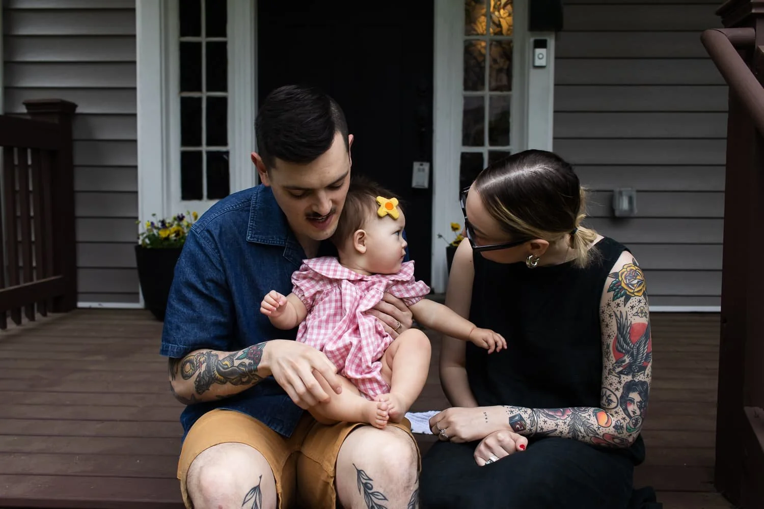 A man, woman, and baby girl sitting on a wooden porch outside a house, sharing a moment of affection and interaction.