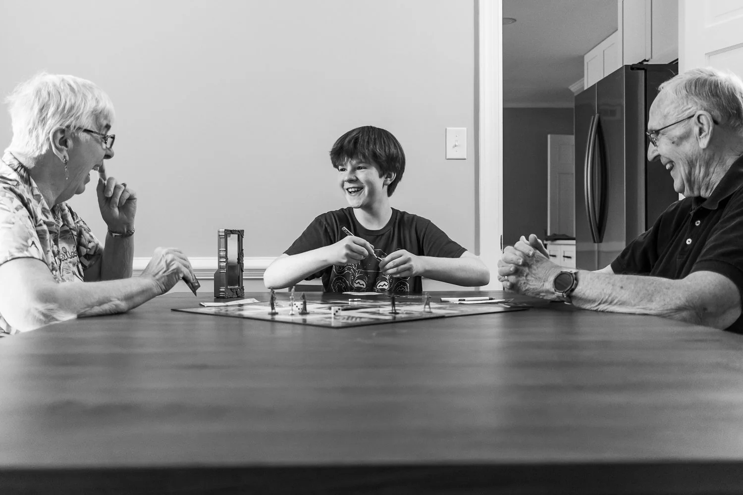 Three people, an elderly woman, a young boy, and an elderly man, are sitting at a table playing a board game together. They are all smiling and appear to be enjoying themselves.