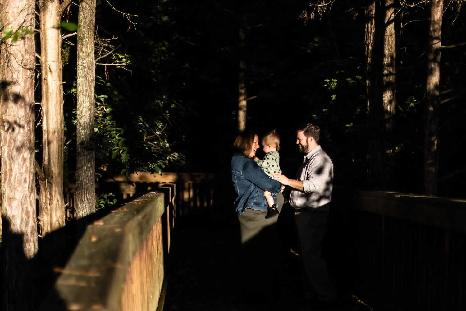A family of three, including a woman, a man, and a young child, are standing on a wooden bridge in a forest at sunset, smiling and interacting joyfully.
