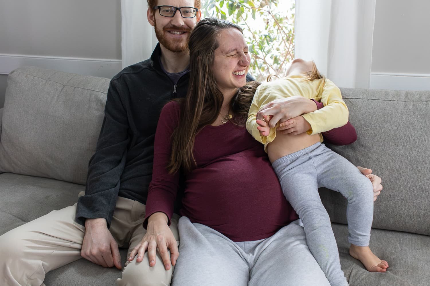 A couple with a young girl on a gray couch, laughing and playing together in a bright room with natural light and curtains.