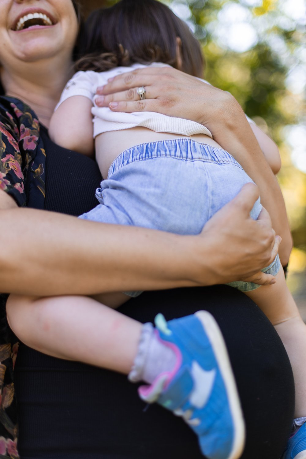 A woman holding a young child outdoors, with part of her face visible showing a big smile. The child is wearing a white shirt, blue shorts, and colorful sneakers, and is being hugged closely.