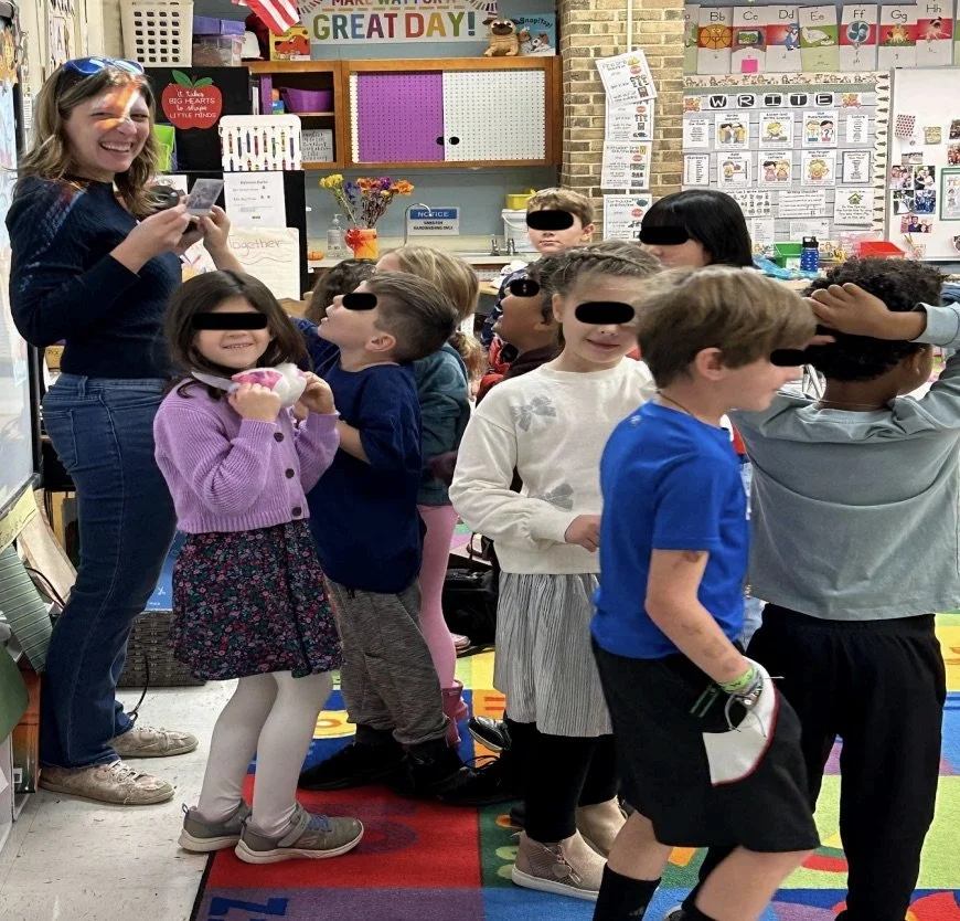A woman stands at the front of a classroom surrounded by a diverse group of 1st graders who all want to hold the Polaroid she's holding in her hand. She's laughing toward the camera. The kids' eyes are blacked out for privacy.