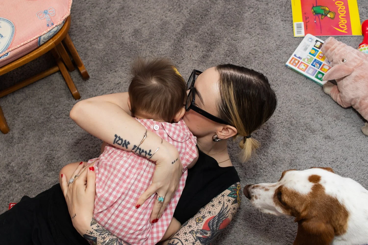 A tattooed mom wearing glasses lays on the carpet and snuggles her baby girl in her arms. The floor has books and toys around her, and the dog is close by.