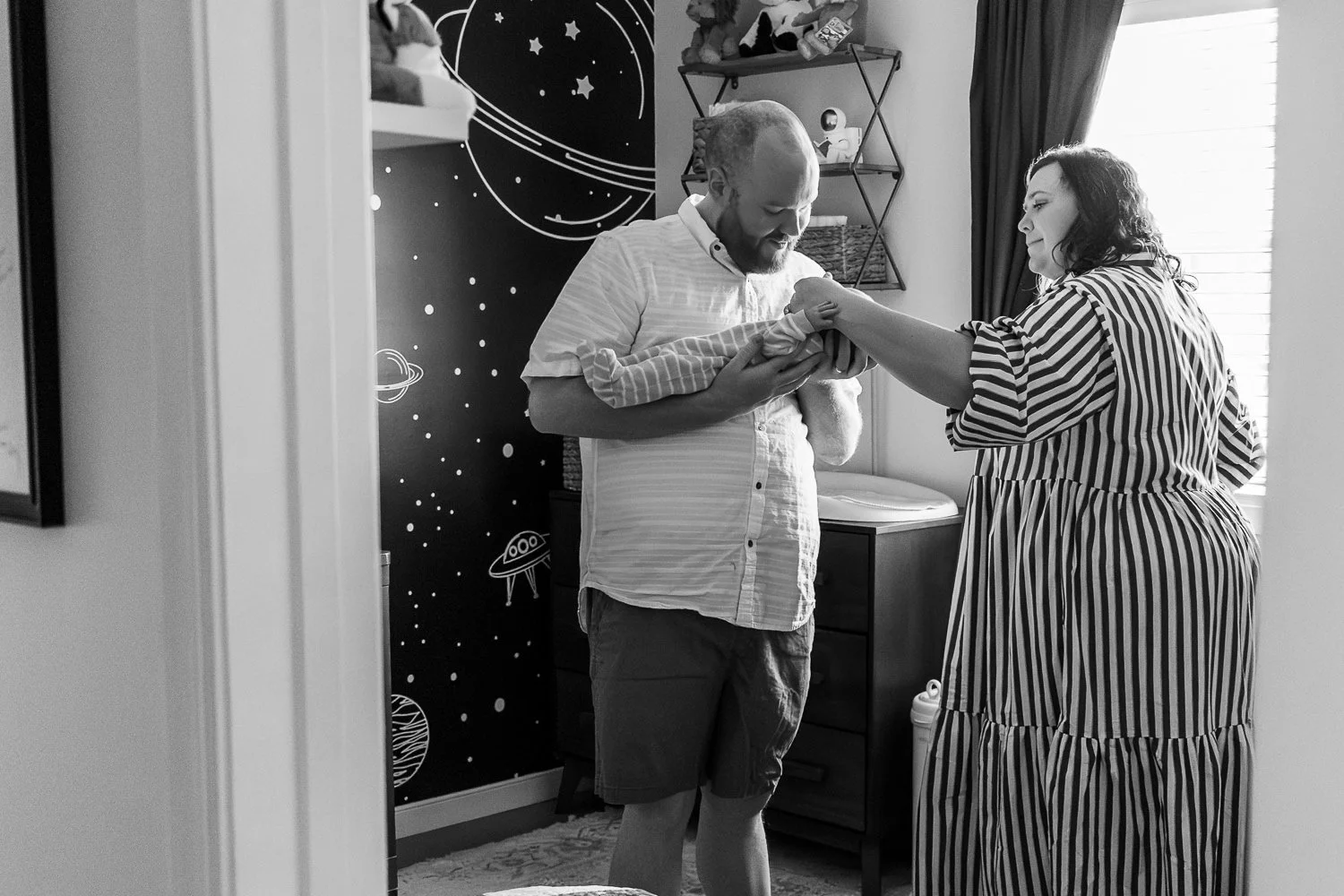 A family of three standing in the baby's nursery. Dad is holding baby, Mom is reaching in to comfort the baby, and baby is reaching up and grabbing mom's arm.