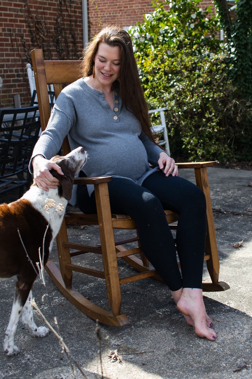 A pregnant woman sits barefoot in a rocking chair on her patio in Richmond, VA and rubs her dog's head. She is smiling down at the dog and the dog is leaning toward her. She's wearing stretchy pants and a long casual shirt.