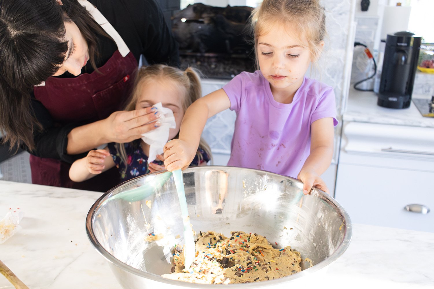A little girl "helps" stir cookie dough in the kitchen at home while her mom wipes her sister's face with a napkin. Both girls are covered in chocolate.