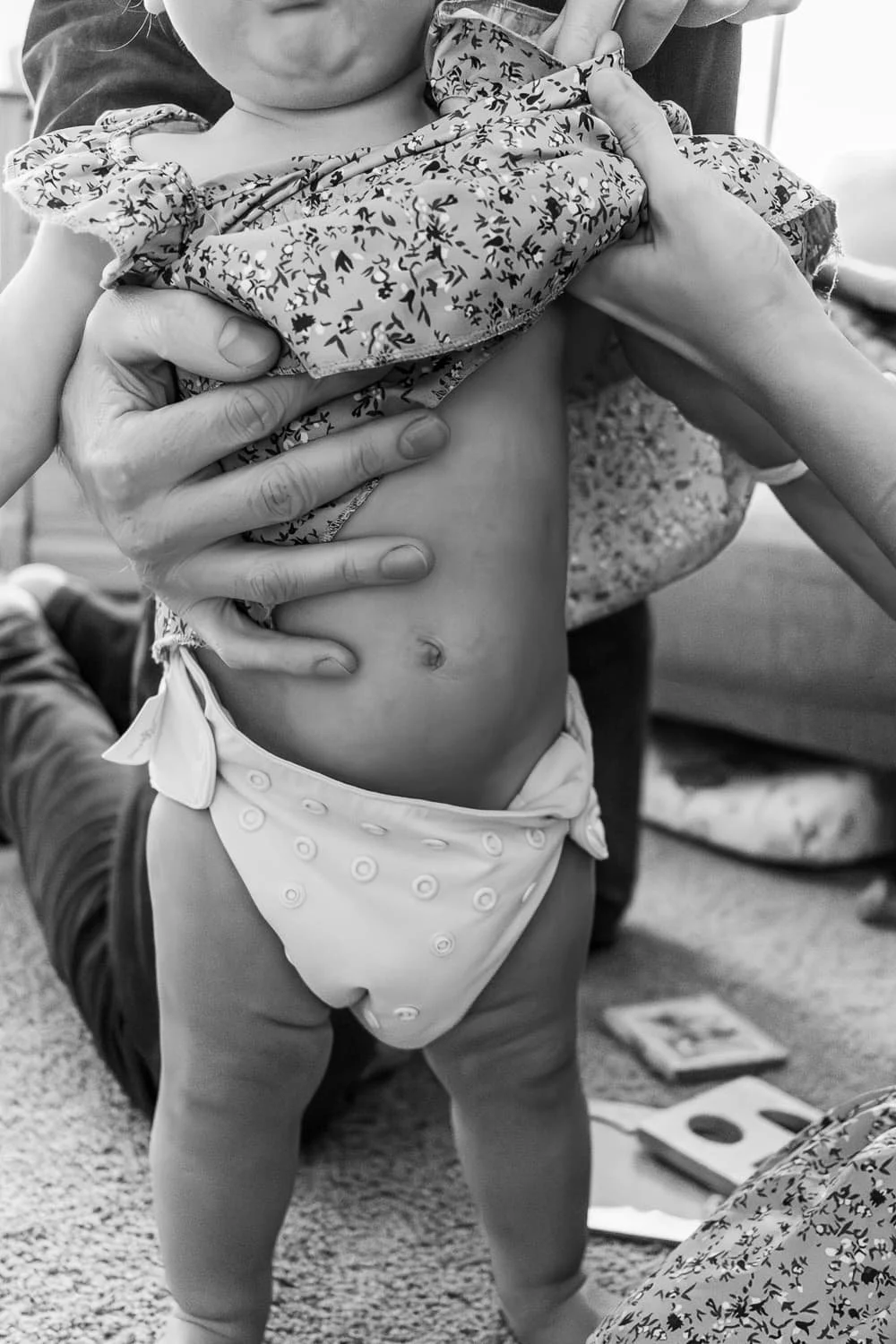 Two parents work together to change the clothes of a standing infant. Her diaper is drooping a little and her belly button shows.