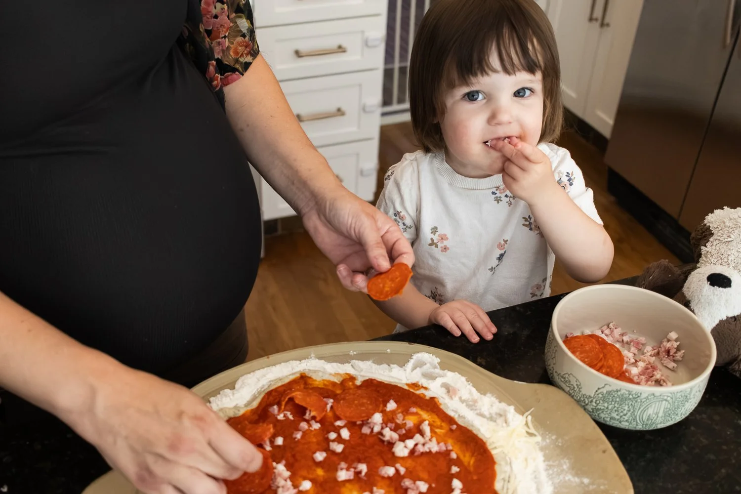 A young girl with brown hair and big blue eyes helping to put toppings on a homemade pizza, with a woman holding a slice of pepperoni, and a bowl of diced ham sitting nearby in a kitchen.