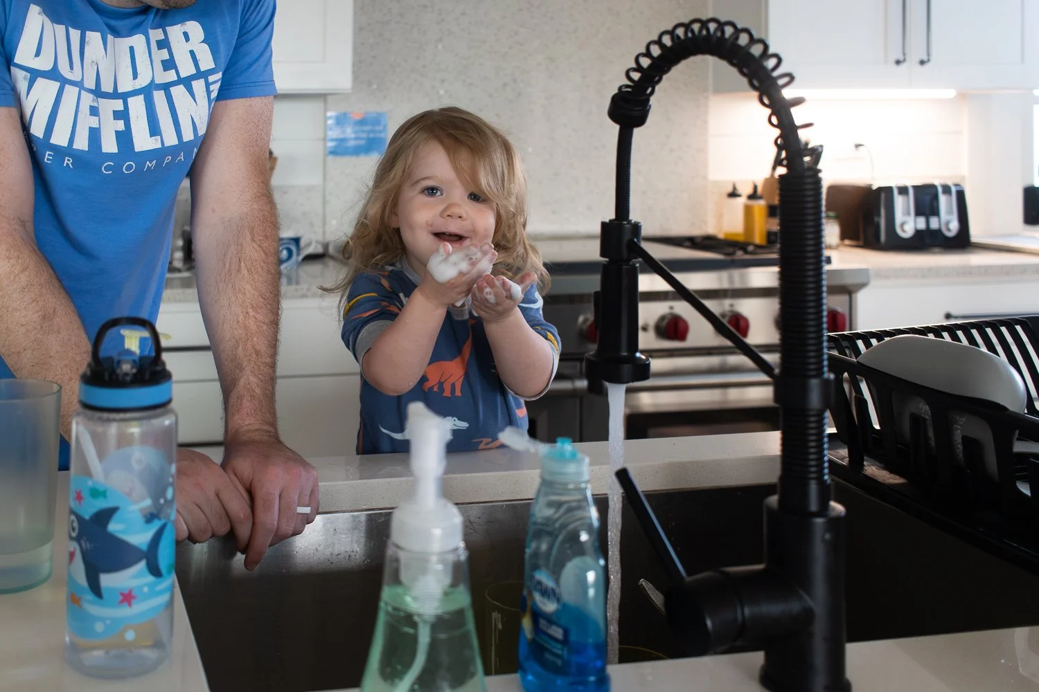 A little girl with soapy hands and a messy face is supervised at the sink by her father. She is smiling, with messy hair and dinosaur pjs.