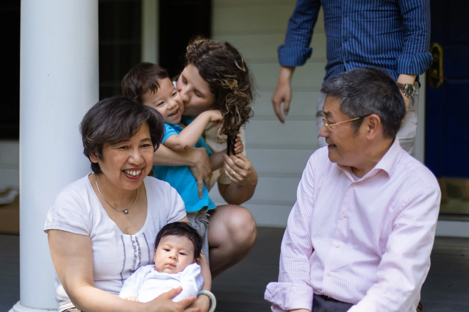Grandparents sit in the foreground on the porch, holding their newborn granddaughter. Mom kisses big brother's cheek behind them while Dad walks into the frame. Everyone is laughing.