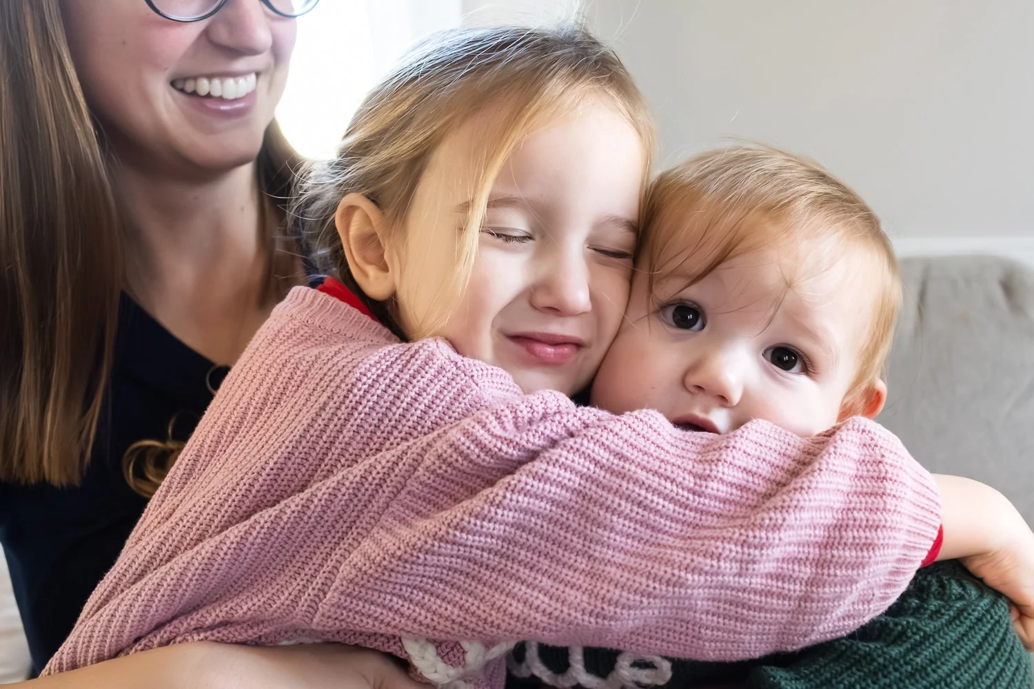 A toddler big sister hugs her infant baby brother and he looks at the camera with pleading eyes as she squeezes her eyes shut