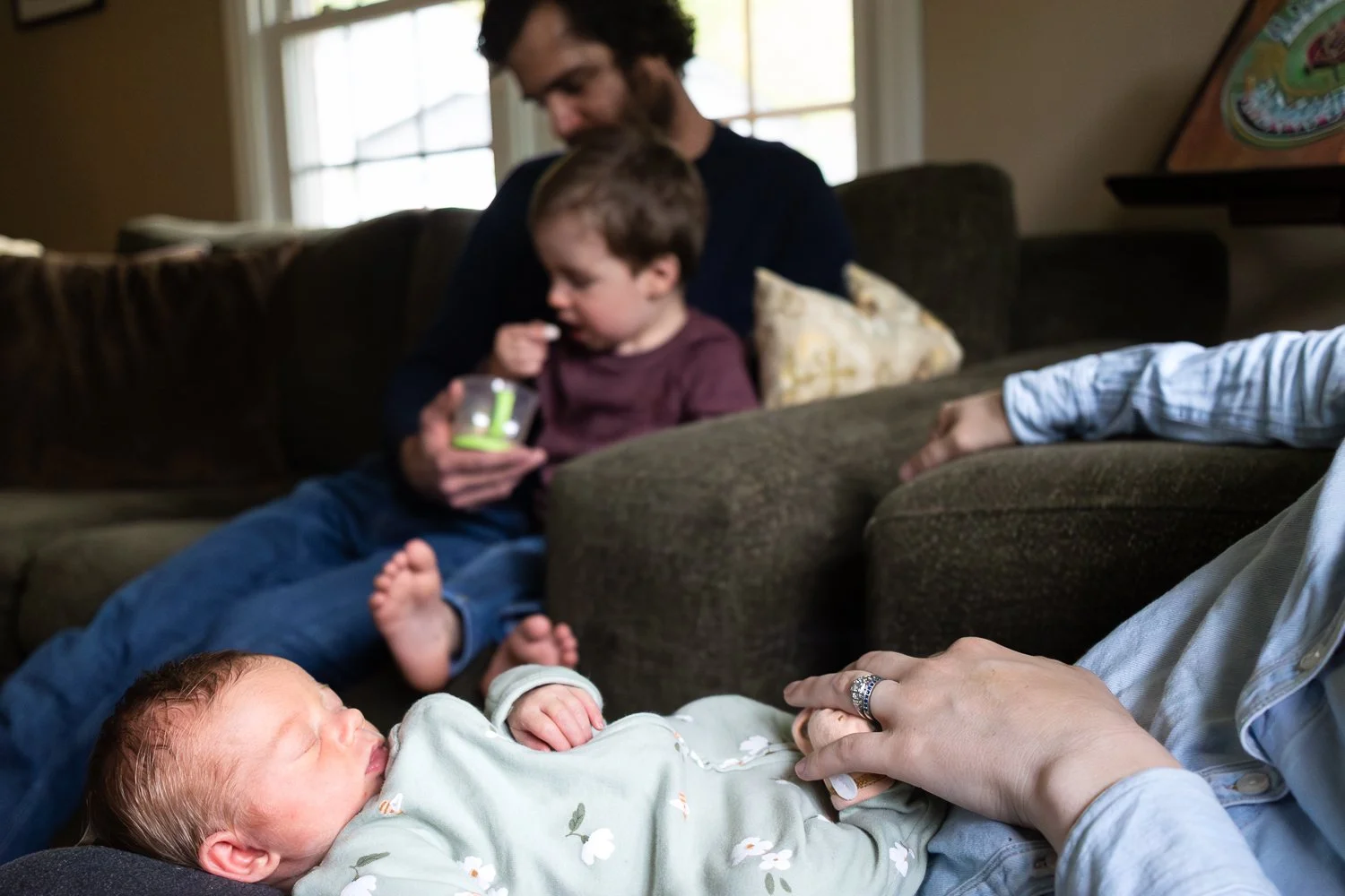 A family relaxes at home during a family photo session in Richmond, VA. Newborn baby sleeps on Mom's lap while big brother eats snacks with Dad in the background.