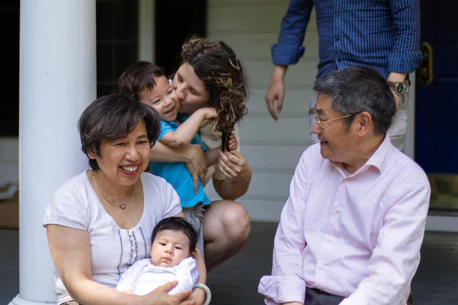 A family gathering on a porch, with a woman holding a baby, a man smiling at her, a woman kissing a young boy, and another person standing behind them.