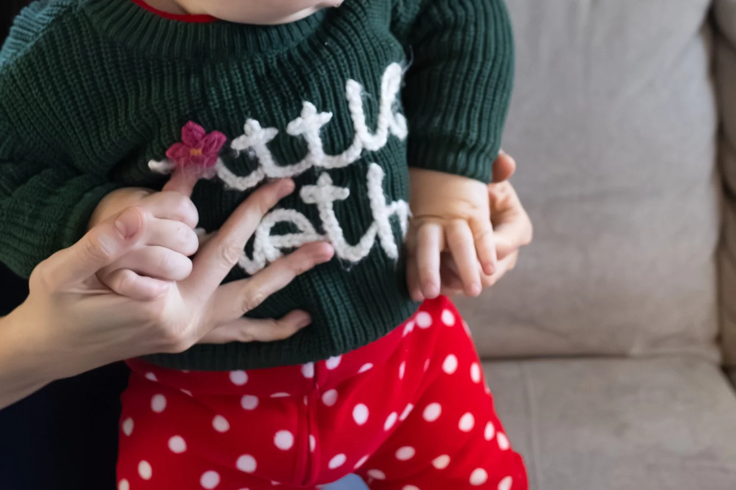 A toddler grabs his mom's fingers as he attempts to stand on the couch at home. He's wearing a sweater that says Little Brother.