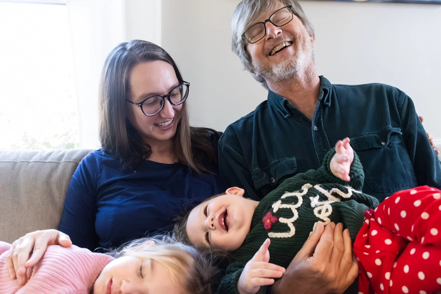 Mom sits on the couch with her dad and her two kids, as they play together. Everyone is playing or acting silly, no one is looking at the camera.