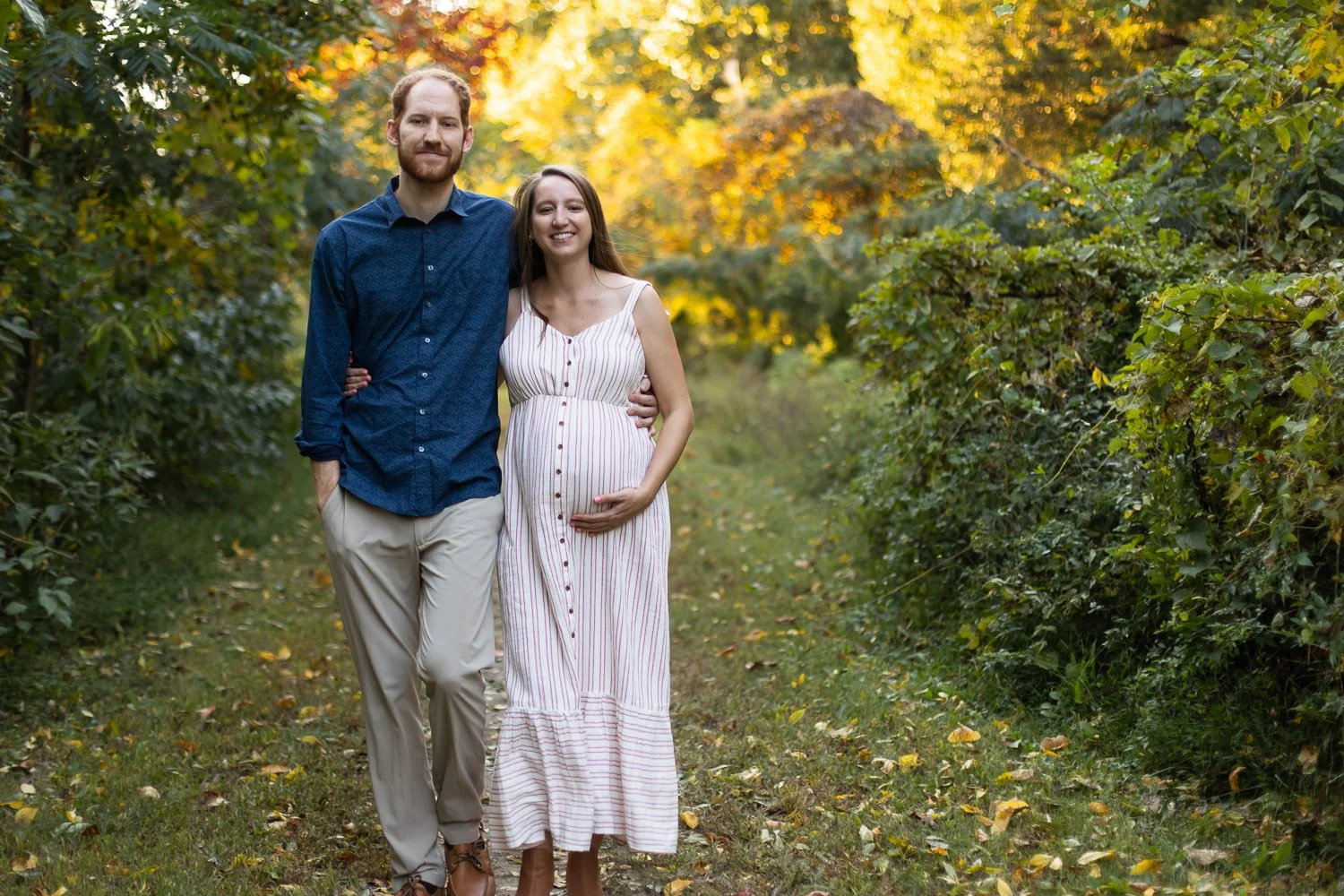 An expecting couple walks along a dirt path at Pumphouse Park in Richmond, VA, looking and walking toward the camera. They have their arms around each other and they're smiling.