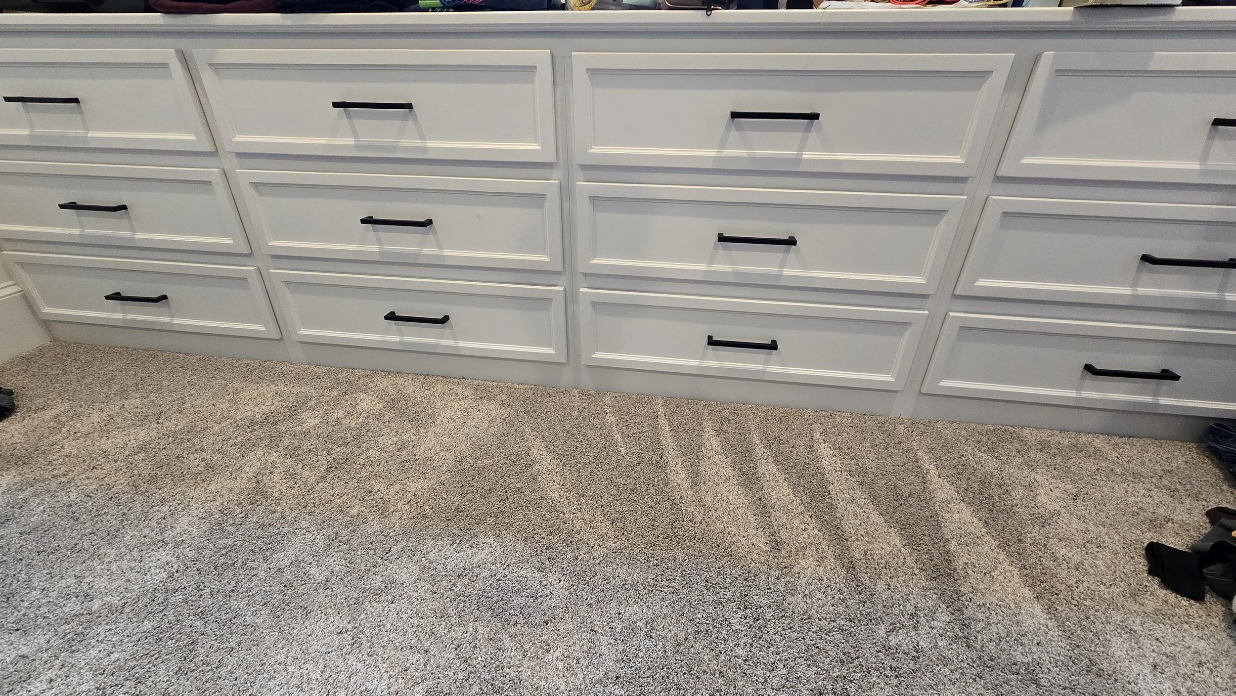 A cellphone picture of 12 white built-in drawers with new black drawer pulls, sitting on top of carpet.