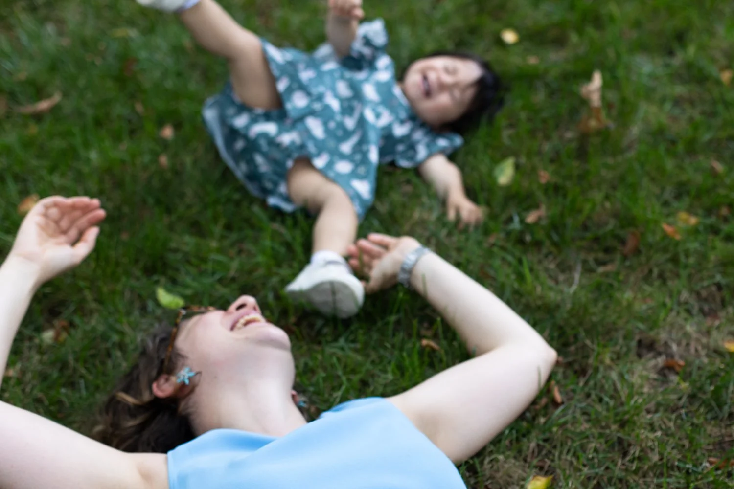 A mom and her toddler daughter laugh while laying in the grass in their yard.