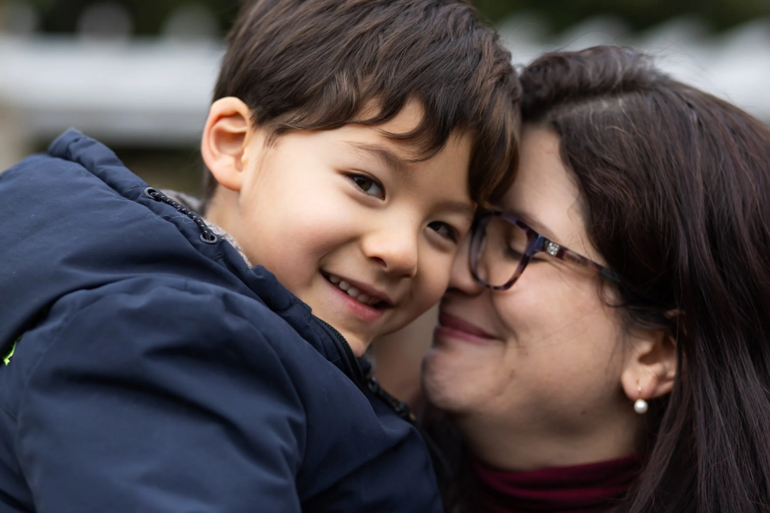 A mom buries her smiling face into her son's cheek while he grins at the camera. They're wearing coats.
