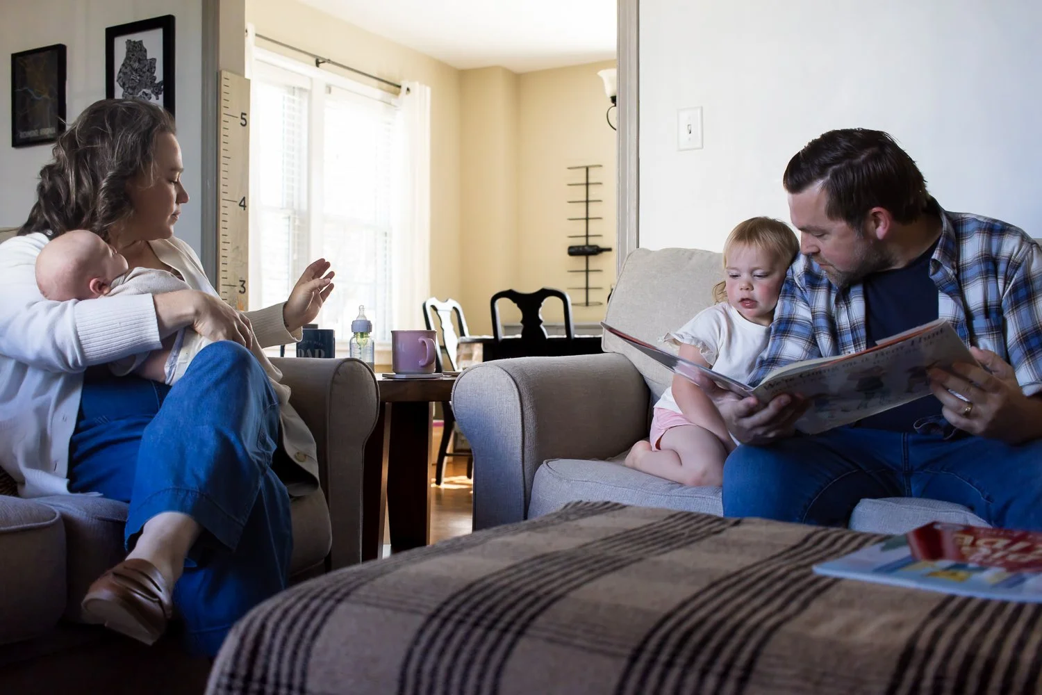 A mom reaches for her baby's bottle on the table beside her while Dad and toddler daughter cuddle up and read a book on the couch nearby. They are calming nervous systems.