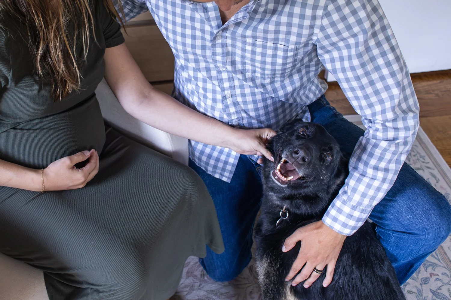 A couple and a black dog sitting on a rug in a cozy living space, with the woman wearing a green dress with a pregnant belly, and the man wearing a checkered shirt. The dog appears happy, looking up with its mouth open.