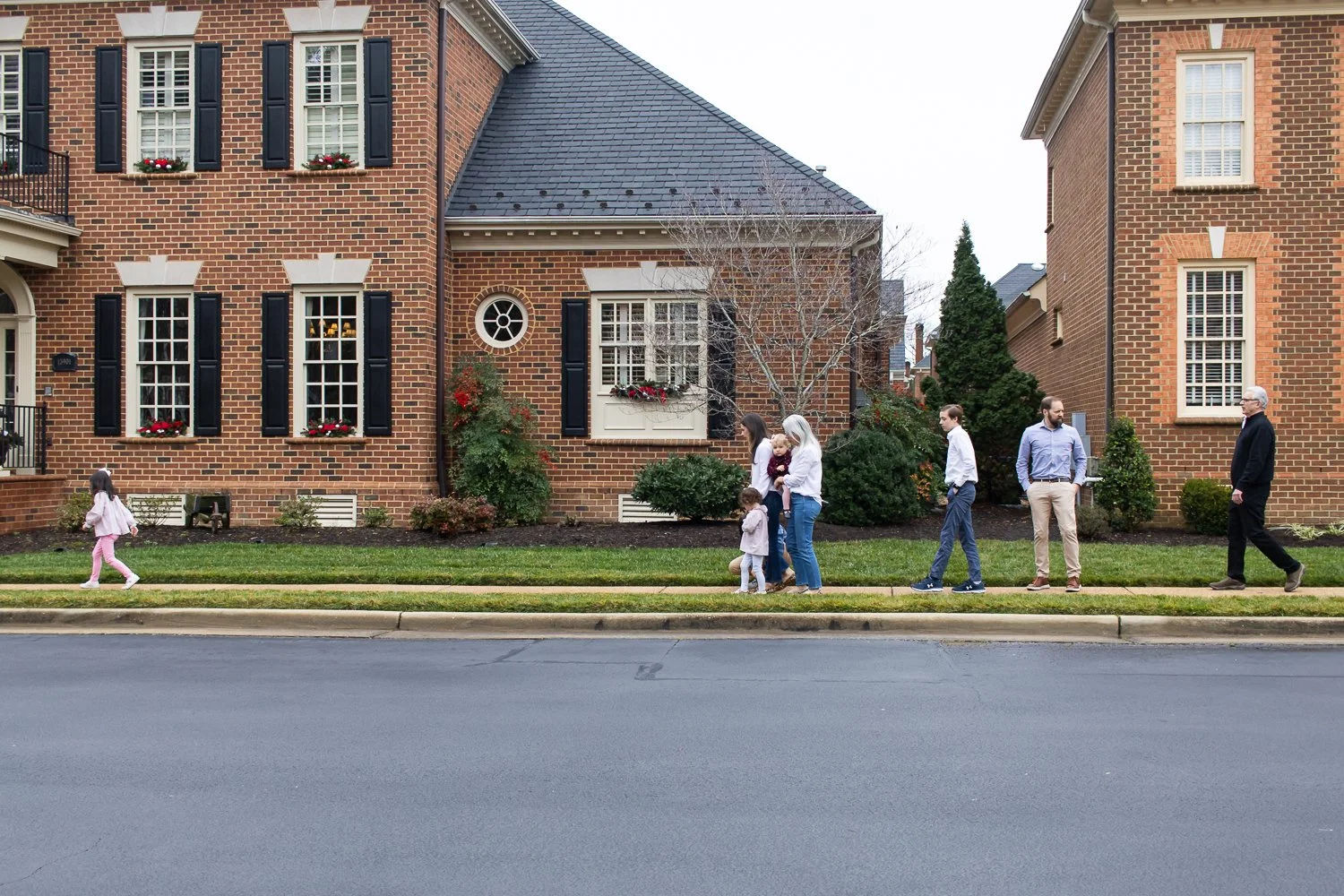 A family walking on the sidewalk in front of brick houses. One girl in pink moves in the foreground, while a group of adults and children walk together in the background. The houses have black shutters, several windows with flower boxes, and some bus