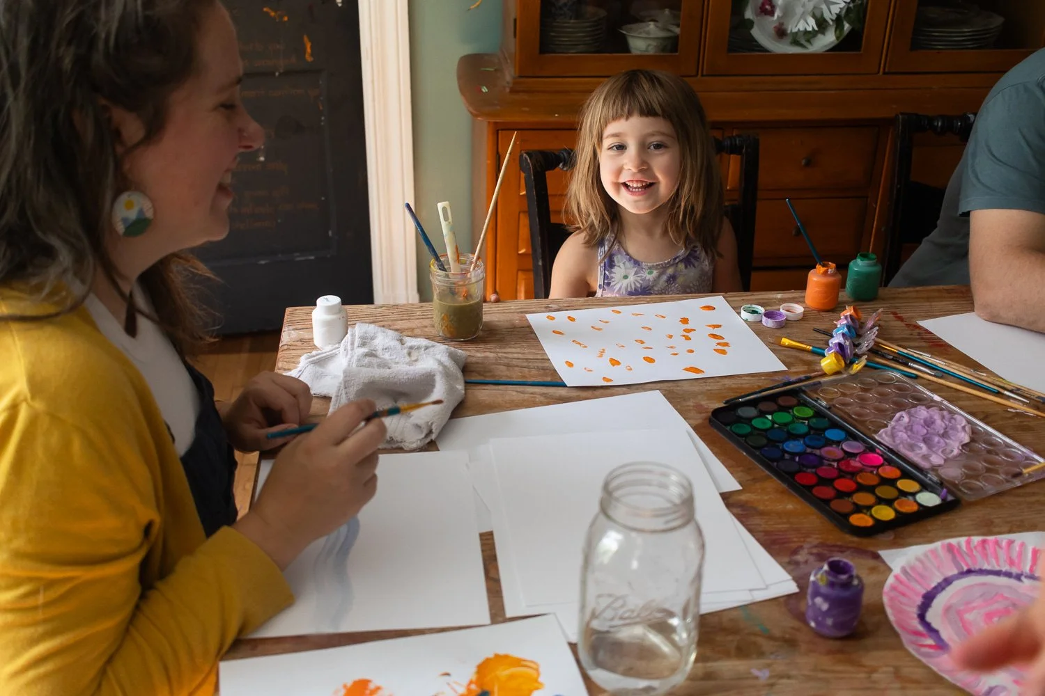 A little girl laughs at the camera with artwork spread in front of her. Her mom is laughing at the table with her own art.