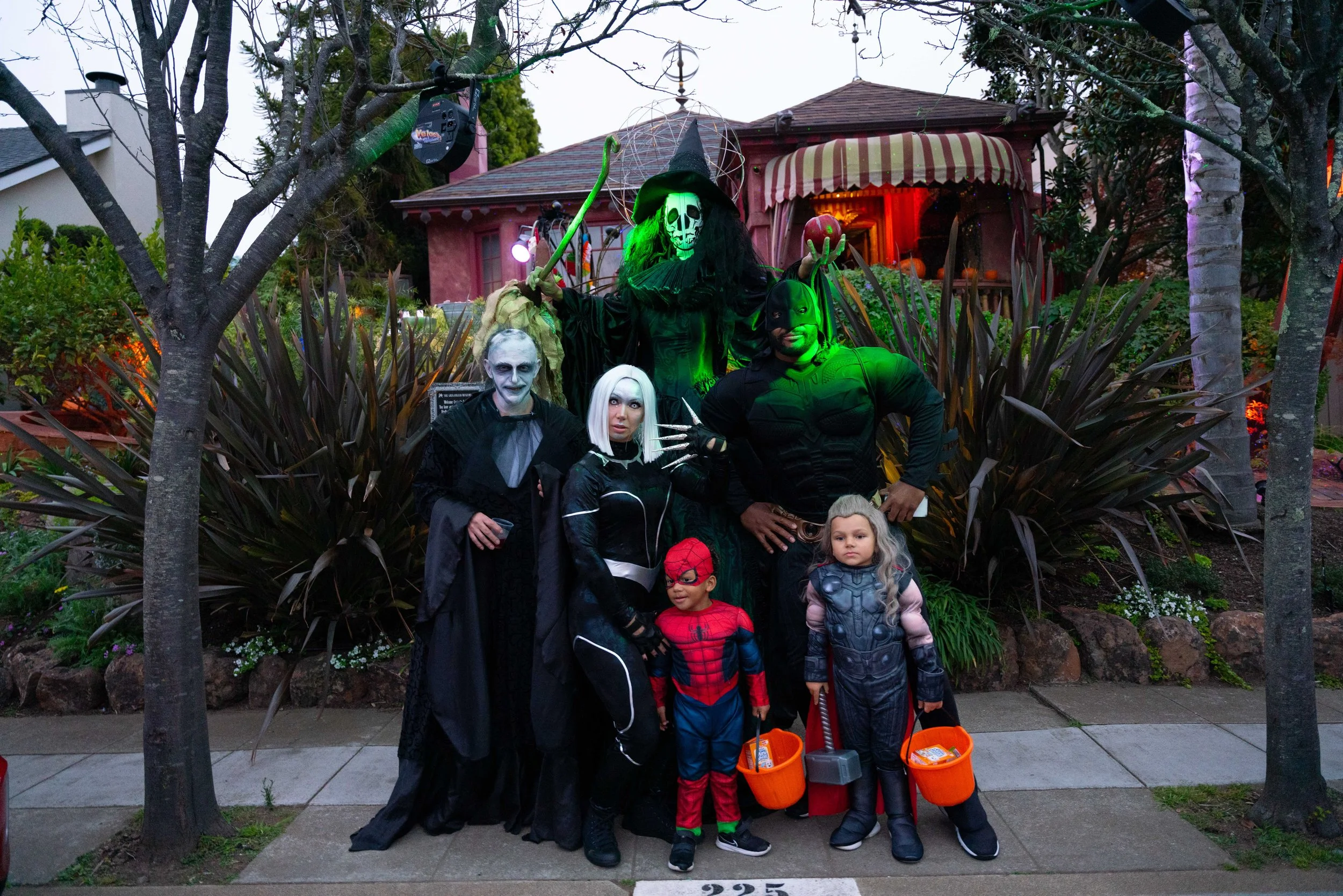 People dressed in Halloween costumes standing on a sidewalk in front of Halloween decorations, including a large skeleton figure and spooky house, during dusk.