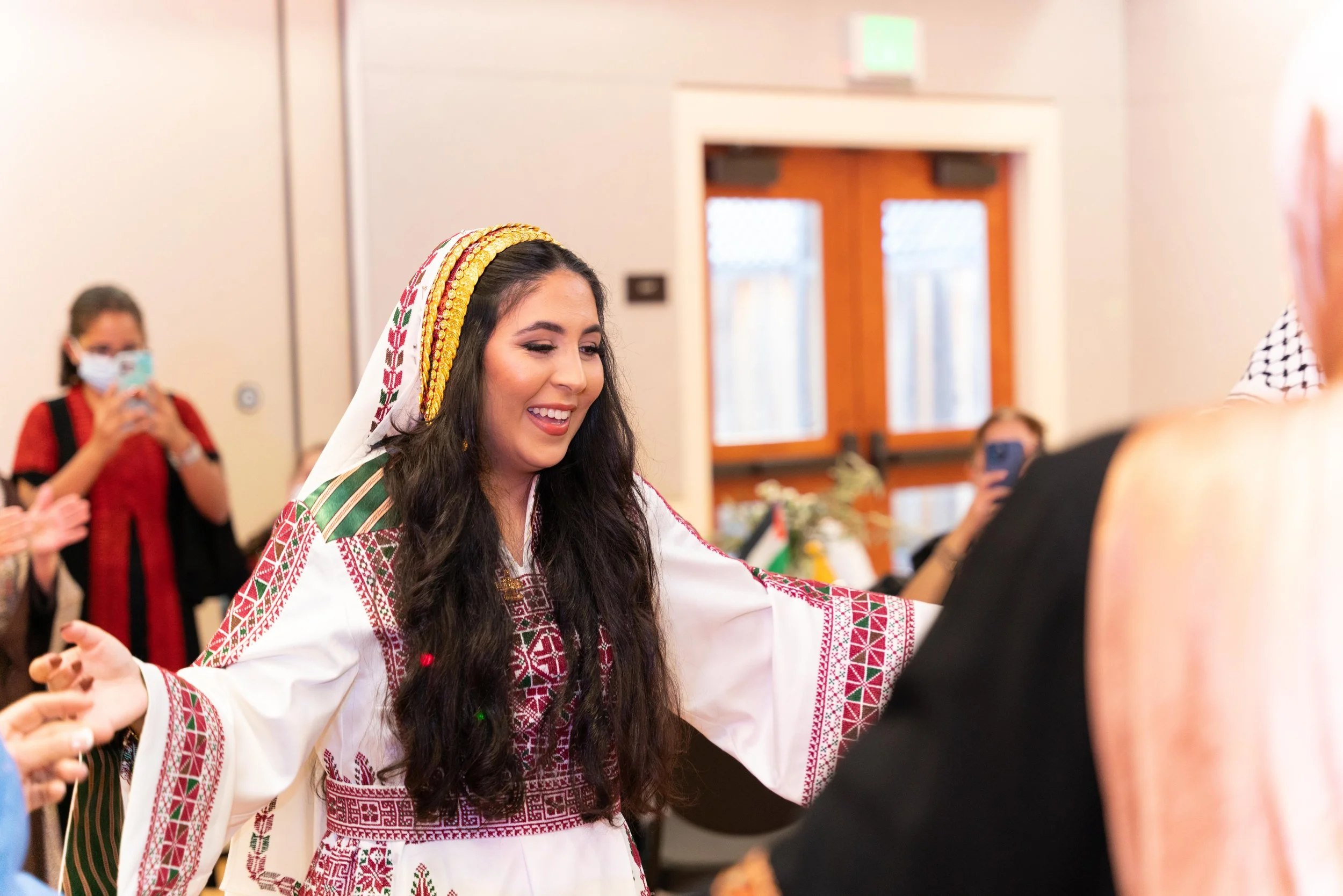 A woman dressed in traditional Palestinian embroidery clothing, smiling with arms outstretched in a celebration or dance, surrounded by other people, some taking photos.