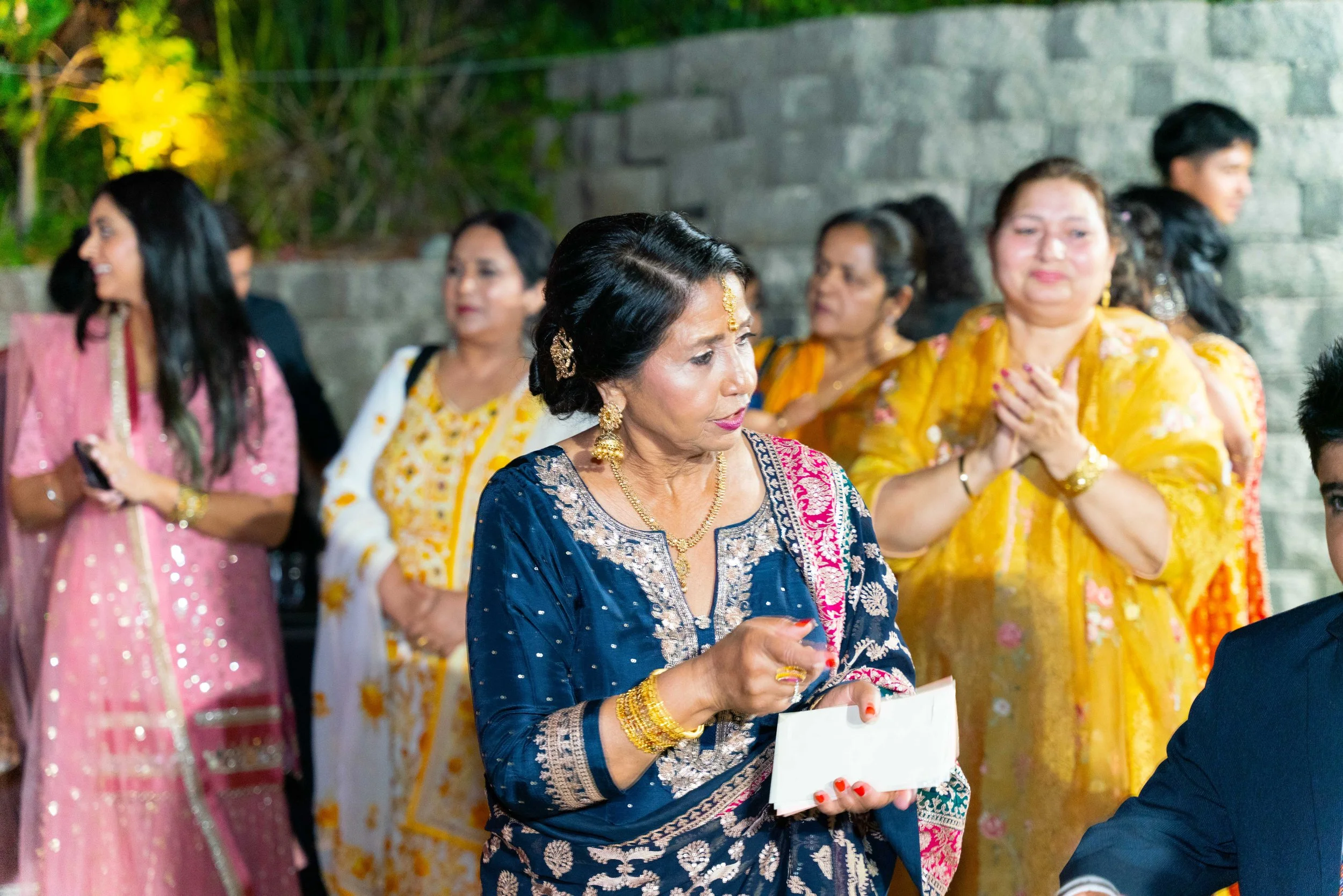 A group of women celebrating at a cultural event, dressed in colorful traditional Indian attire, with some holding smartphones and others clasping hands in prayer or greeting. The central woman wears a navy blue and gold embroidered outfit, holding a