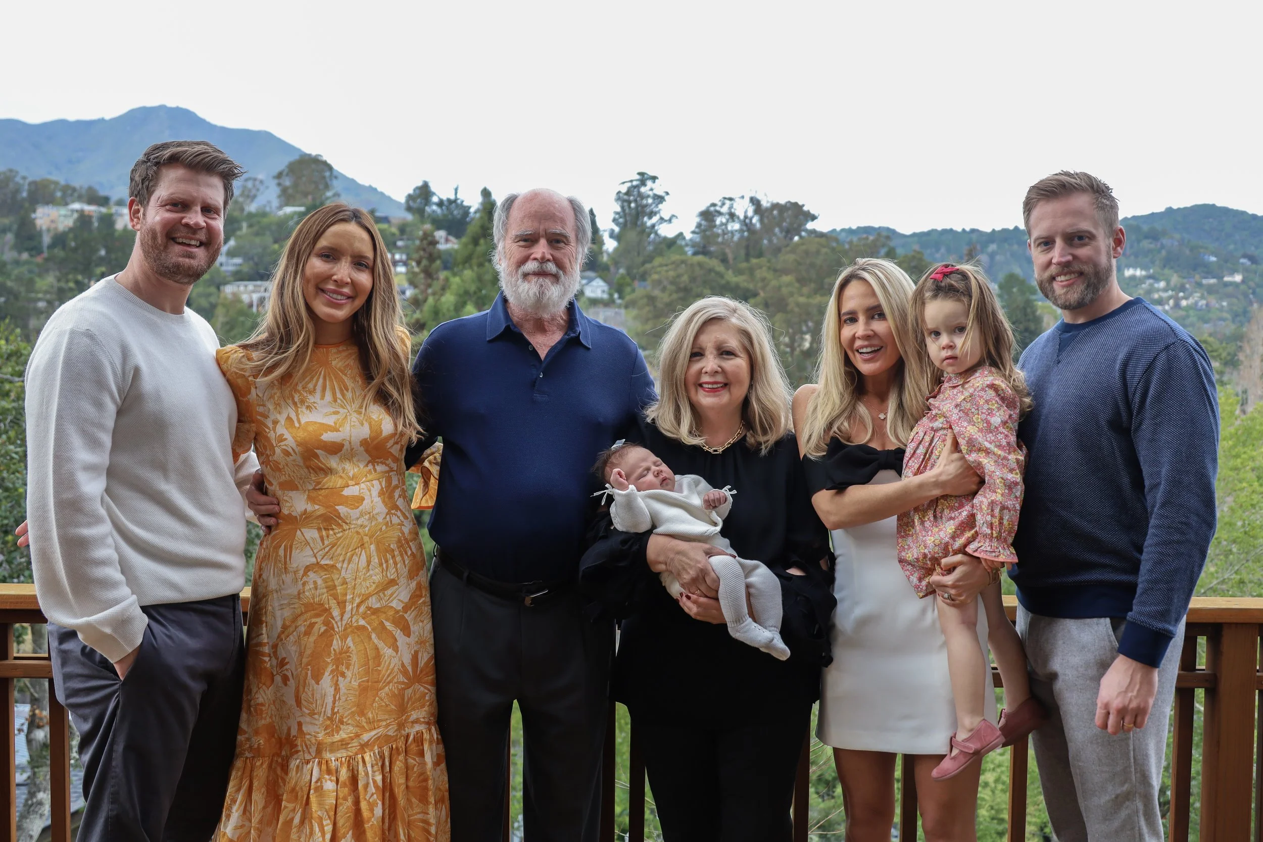 Family group photo outdoors with mountains in the background, featuring a multi-generational family including two children, three women, three men, and a newborn baby.