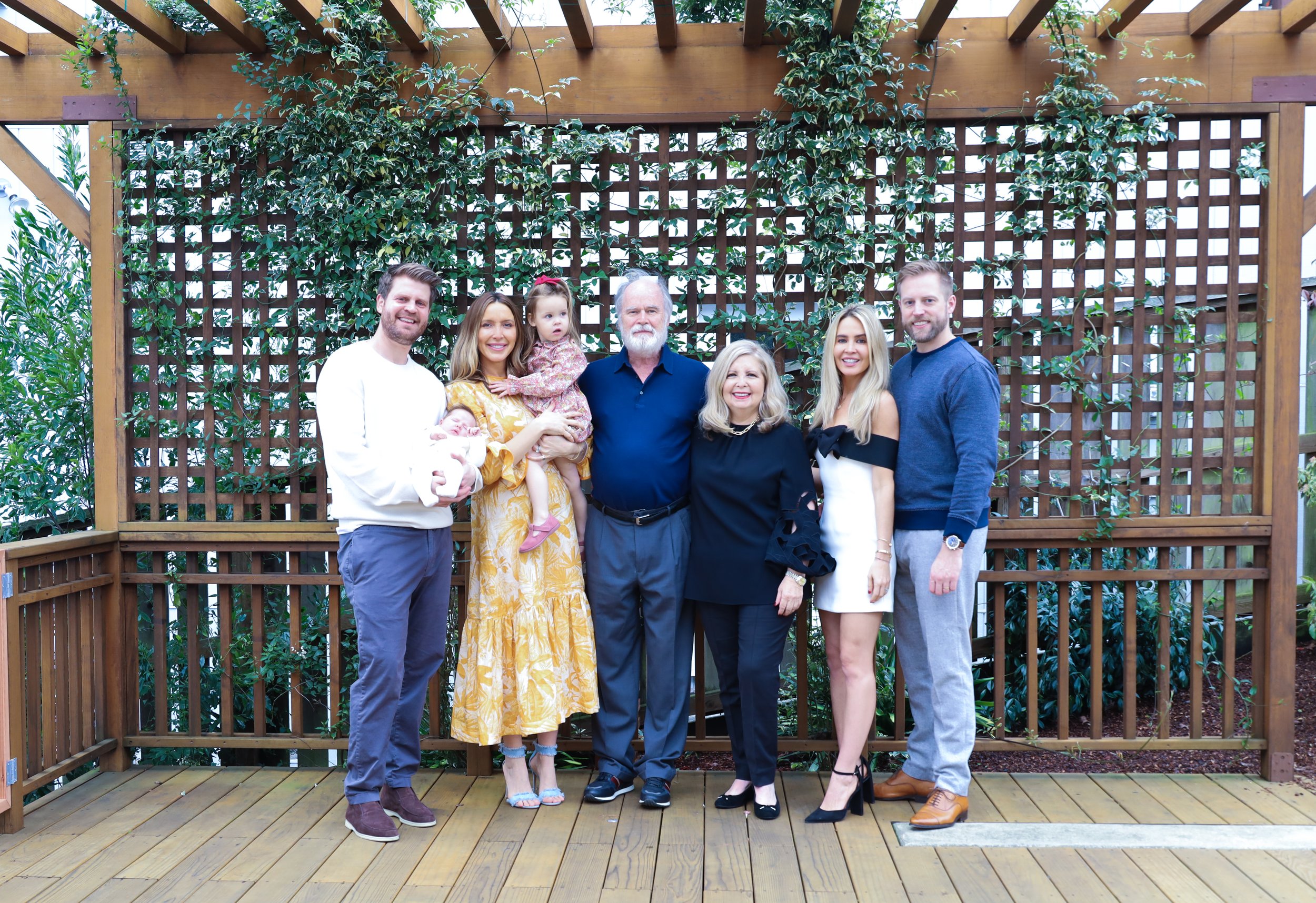 A group of eight people, including four adults, two children and two babies, standing on a wooden deck in front of a lattice wall with climbing plants, smiling for a family photo.