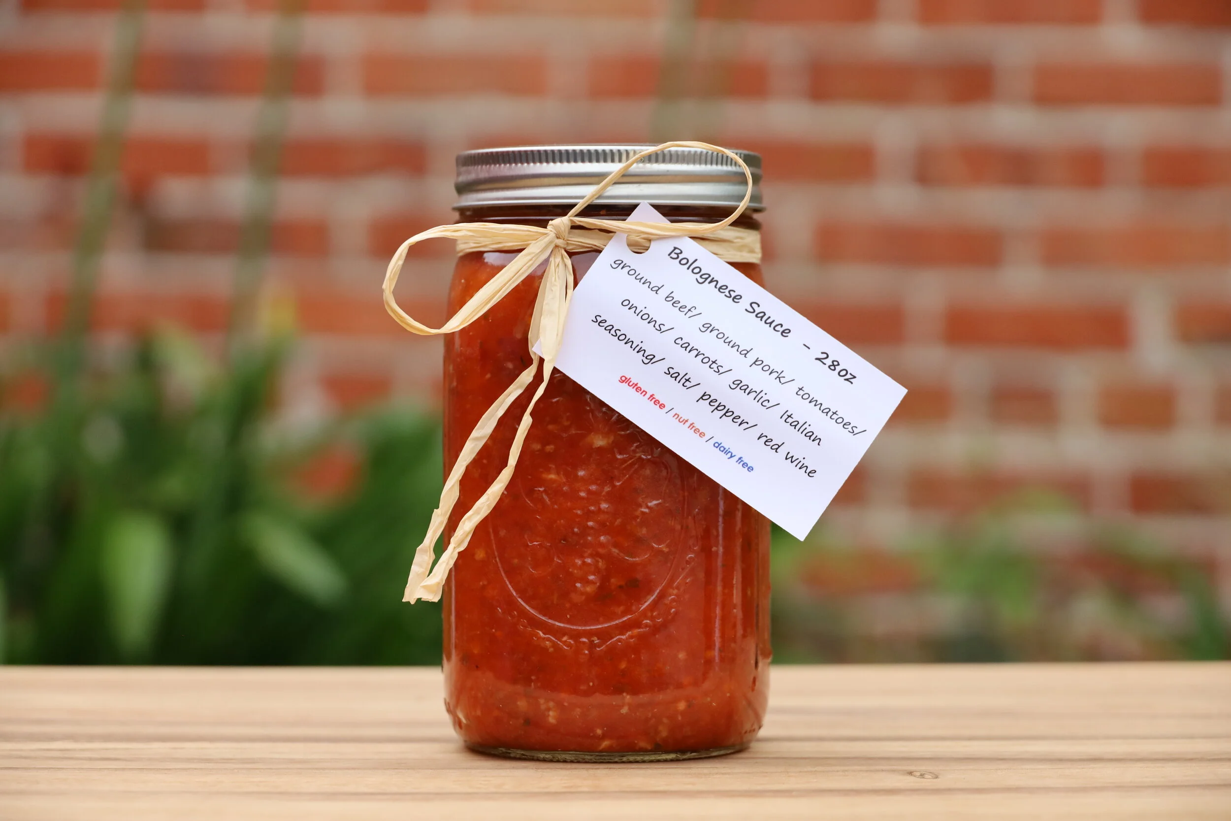 A jar of homemade Bolognese sauce with a silver lid, tied with a beige ribbon. The jar has a white label listing ingredients: ground beef, ground pork, tomatoes, onions, carrots, garlic, Italian seasoning, salt, pepper, and red wine. The background f