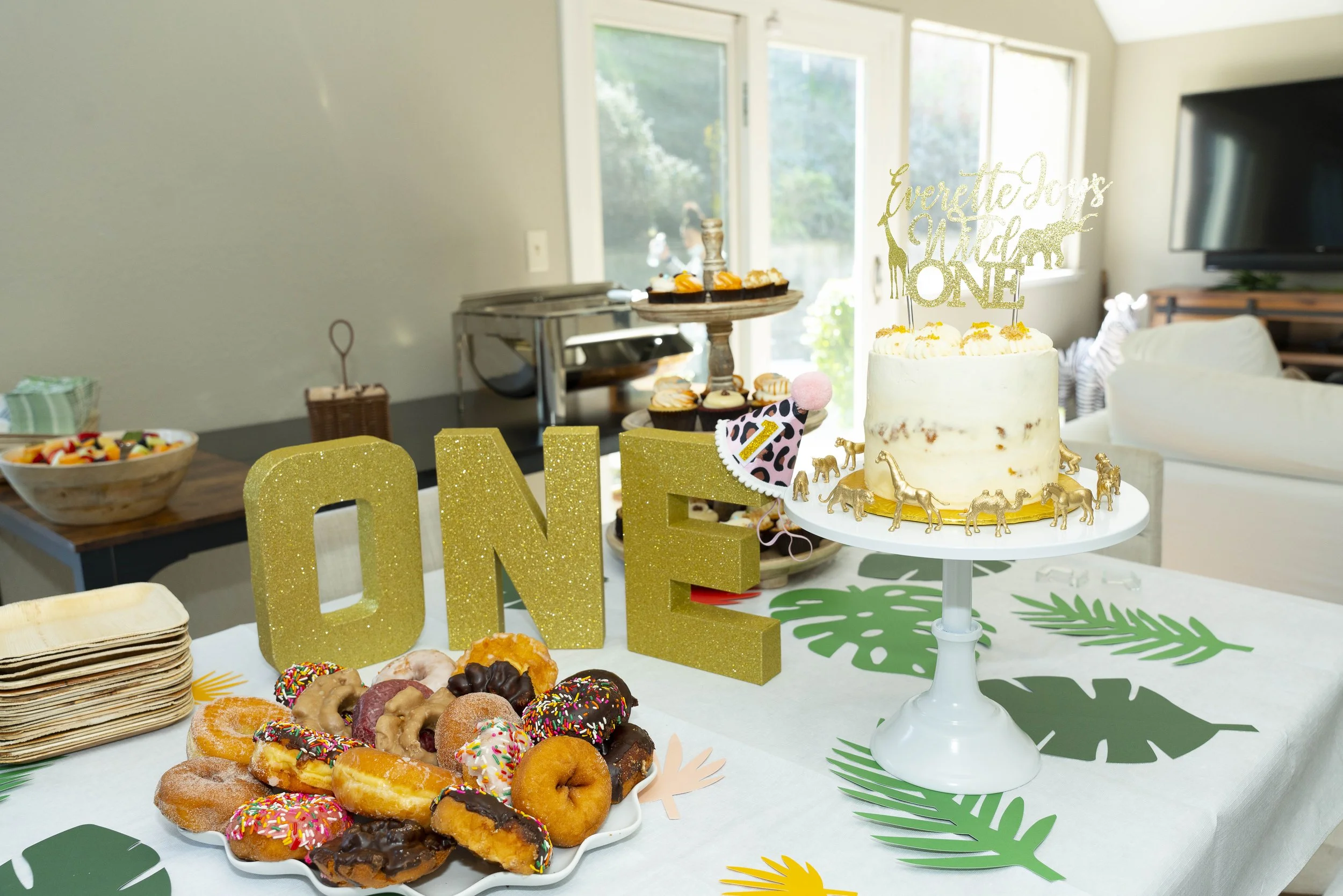Dessert table for a first birthday celebration with a white frosted cake decorated with gold animal figures and a topper that reads 'Everette Jays First ONE,' a variety of colorful donuts with sprinkles, and a gold glitter 'ONE' sign. The table has t