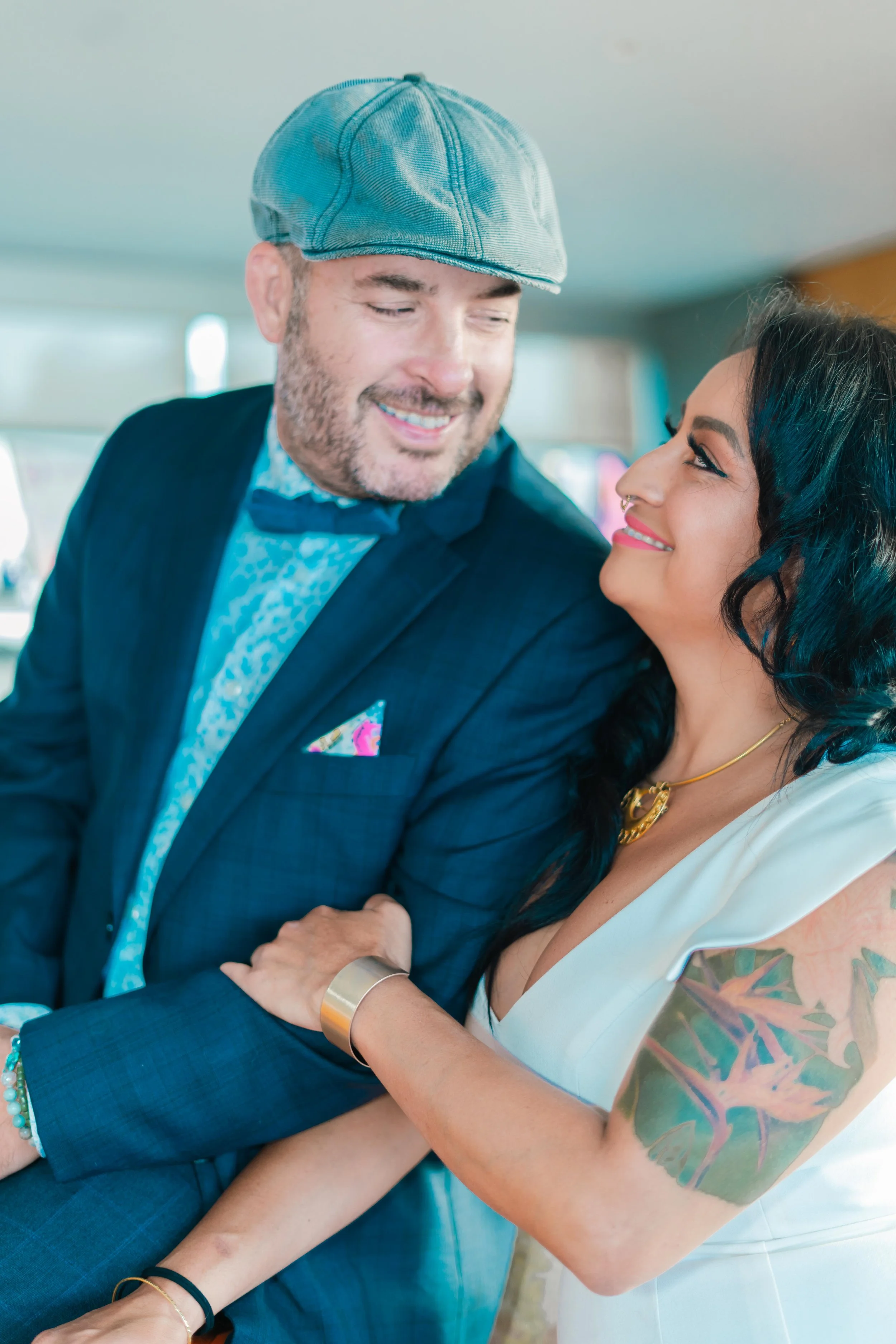 A man and woman gazing into each other's eyes, smiling, with the woman holding the man's arm. The woman has dark hair, tattoos on her arm, and is wearing a white dress and gold jewelry. The man is wearing a blue suit, a blue cap, and a blue shirt wit