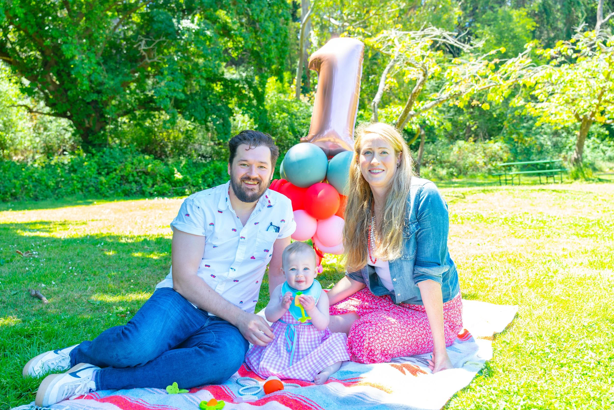 A family of three, with a man, woman, and baby, sitting on a blanket outdoors in a park with trees and grass, celebrating a first birthday with balloons and a large number one balloon.