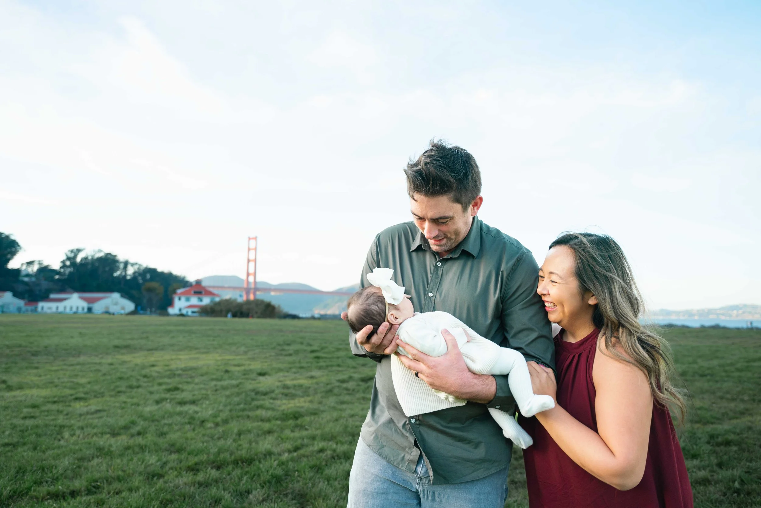 A couple holding a baby outdoors in a grassy park with a bridge and houses in the background, smiling and looking at the baby.